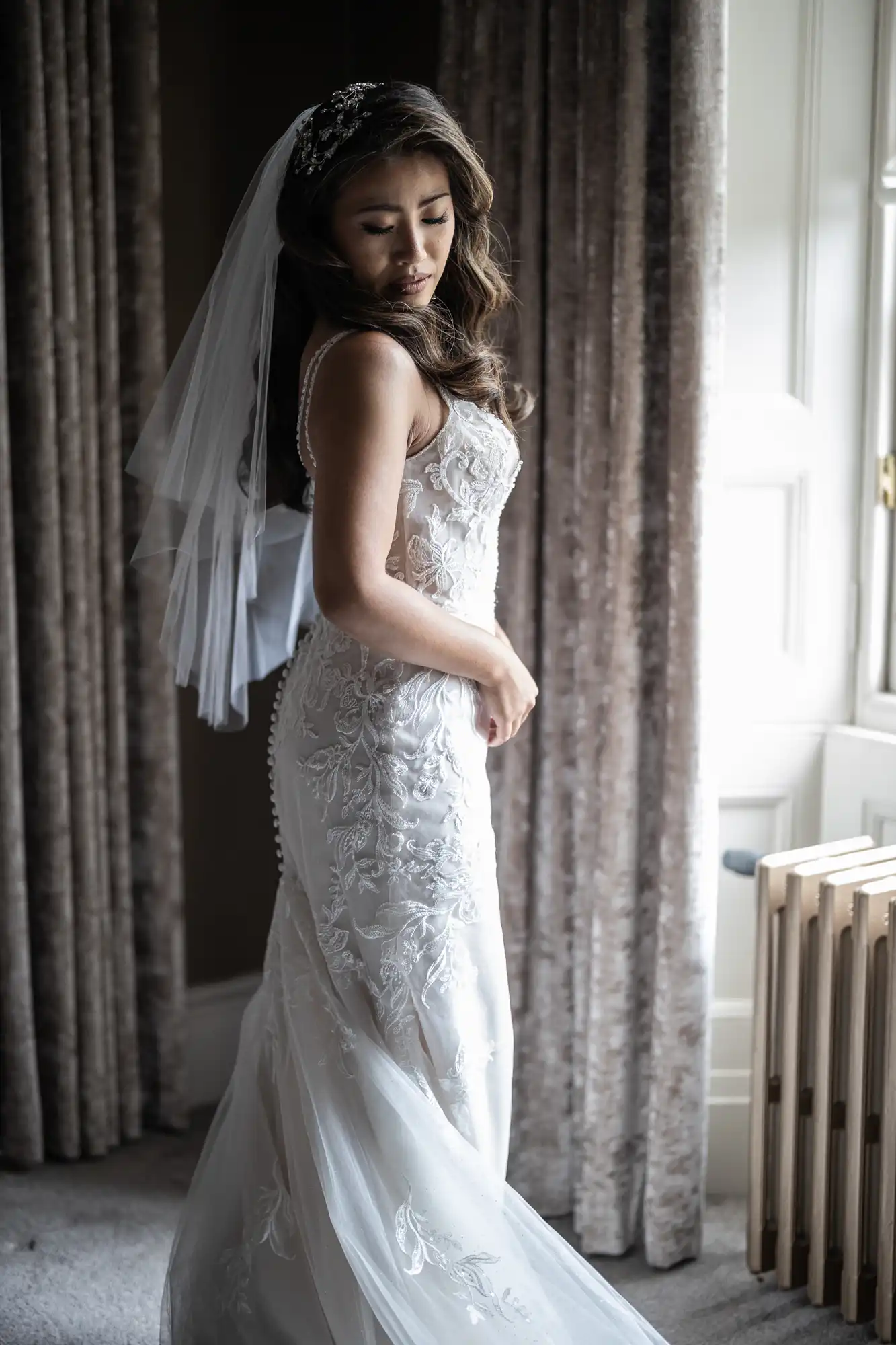 A woman in a detailed white wedding dress stands near a window with curtains, looking down.