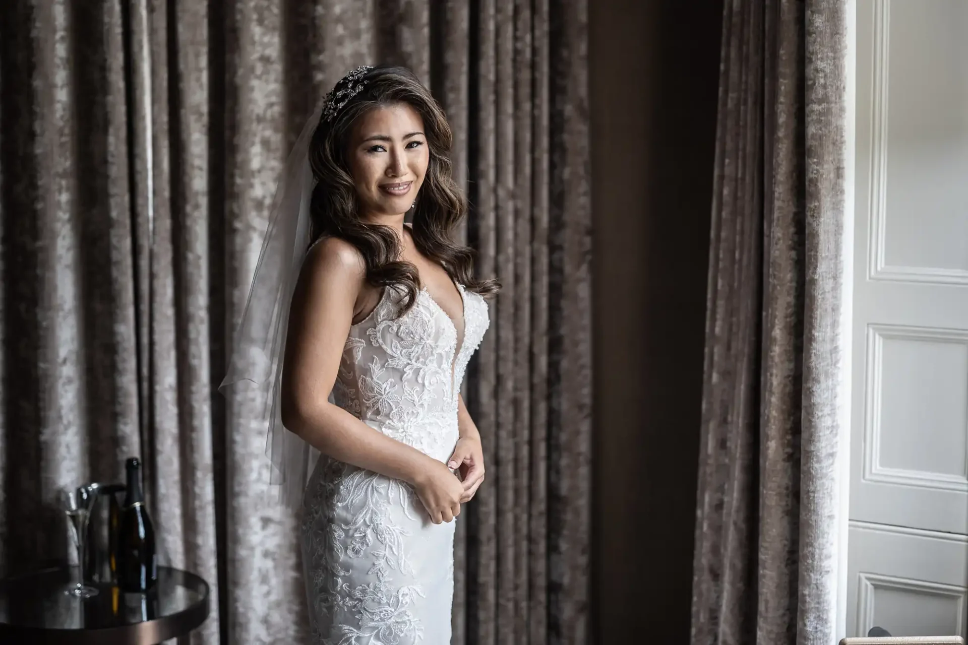 A bride in a white lace dress stands by a window with curtains, smiling. There's a table with a champagne bottle and glasses nearby.