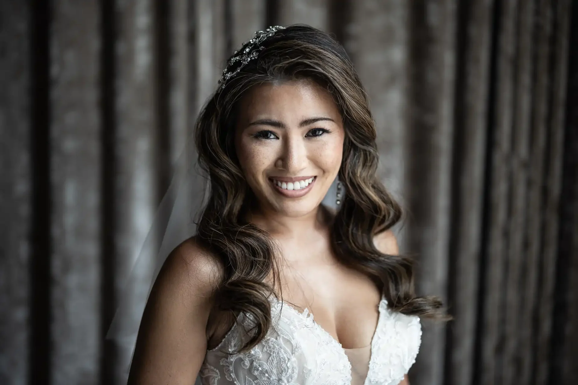 A smiling woman in a wedding dress with wavy hair stands against a curtain background. She wears a beaded hair accessory.