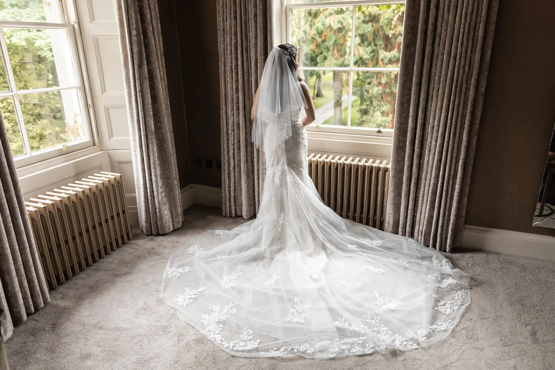 Bridal portrait of a woman in a long, lace-trimmed veil and gown, standing by a large window with heavy curtains, overlooking a garden.