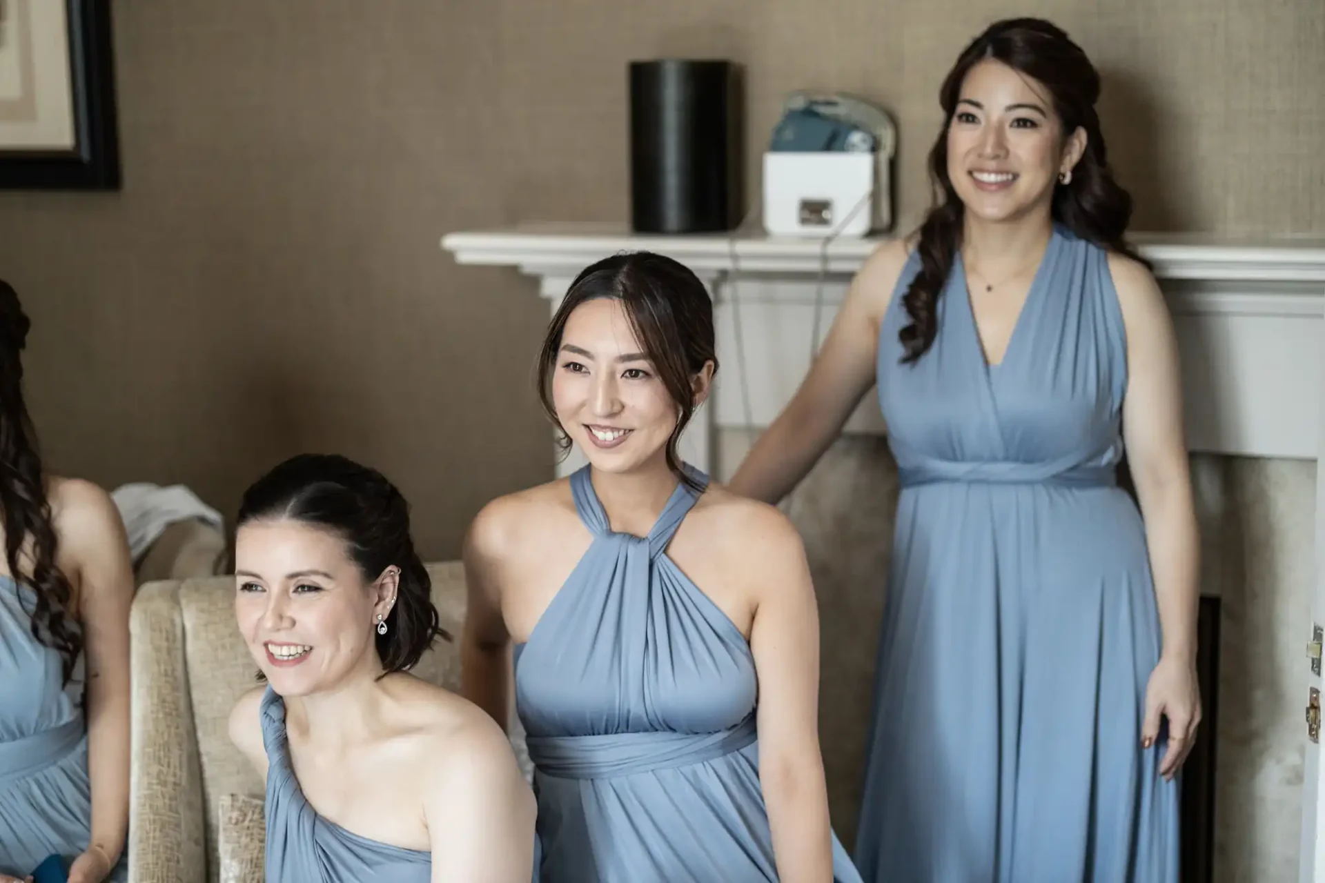 Three women in matching blue dresses smile while sitting and standing in a room with a fireplace.