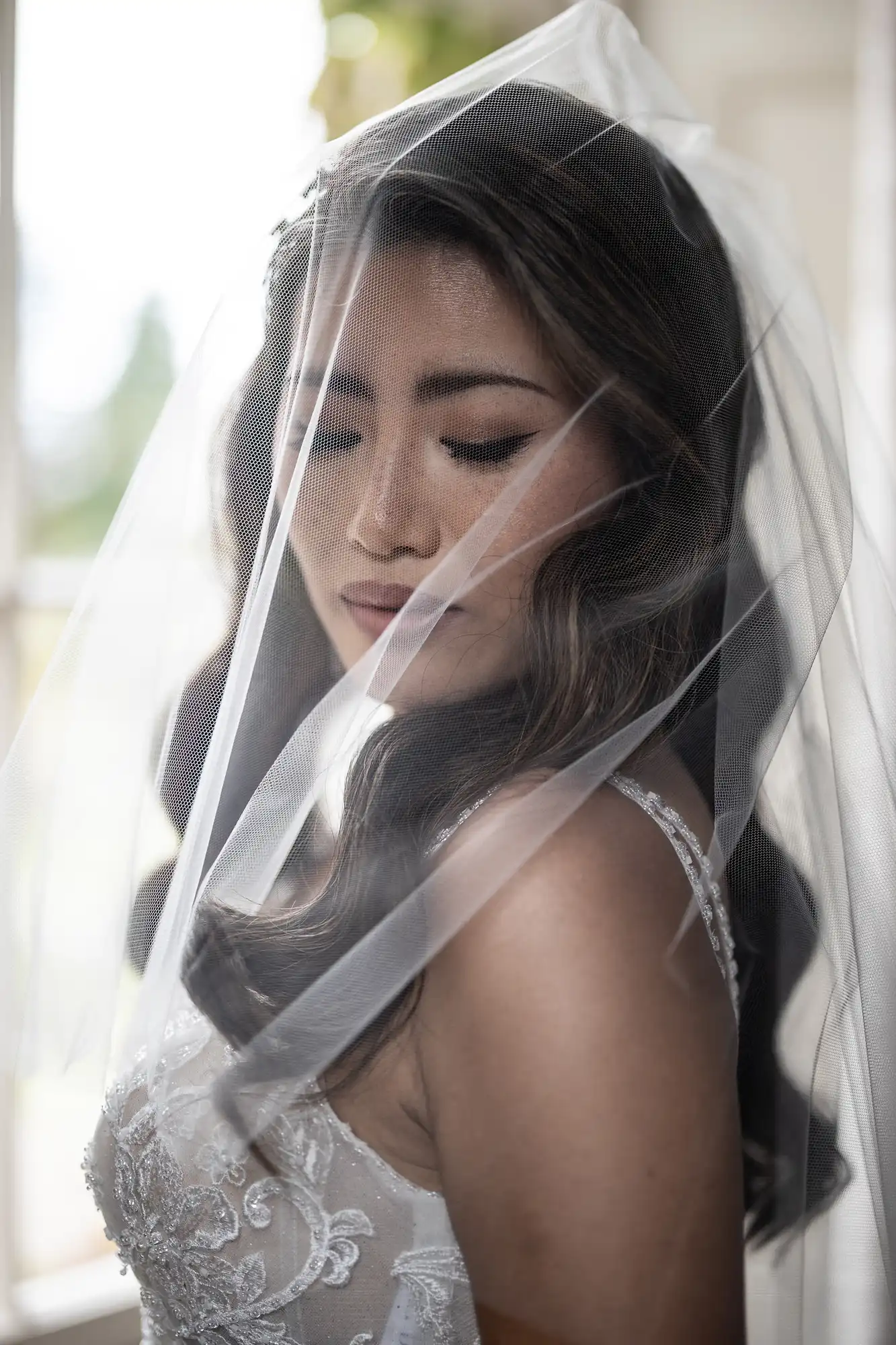 A woman in a wedding dress with a veil looks downward, standing by a bright window.