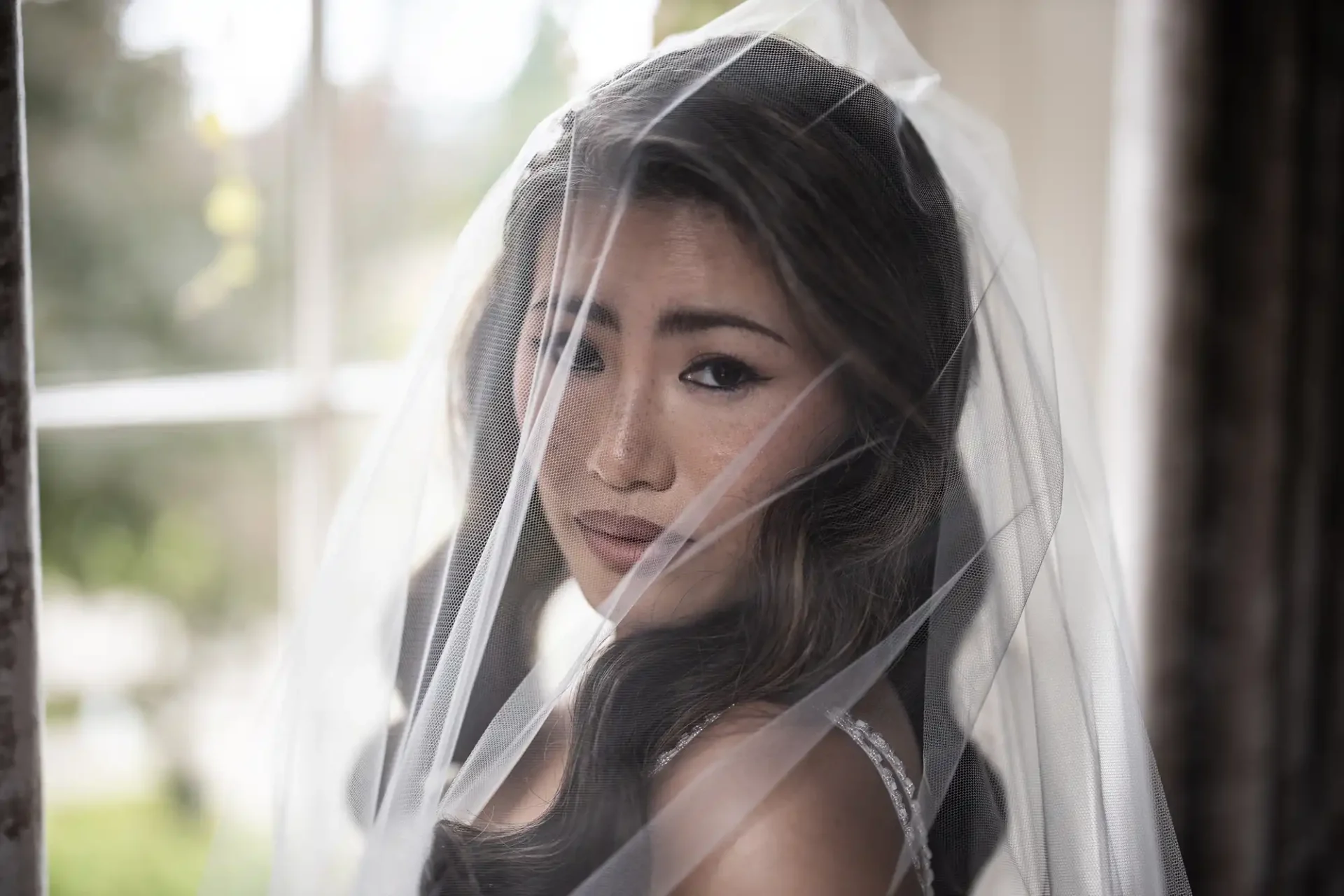 A bride in a sleeveless gown gazes through a sheer veil inside a room with soft lighting.