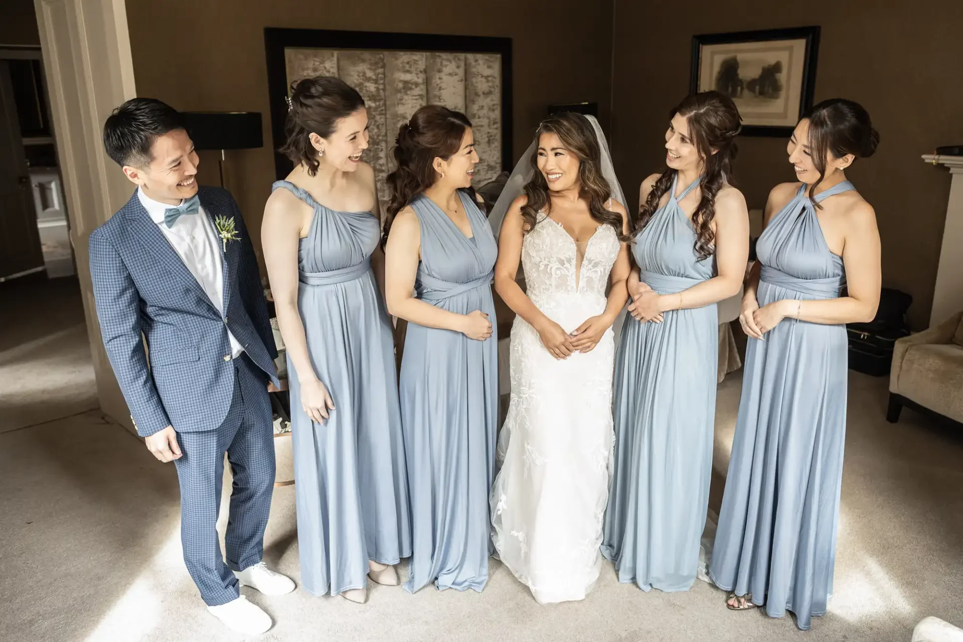 Bride in a white gown stands smiling with five attendants in light blue dresses and one man in a blue suit in a room with muted decor.