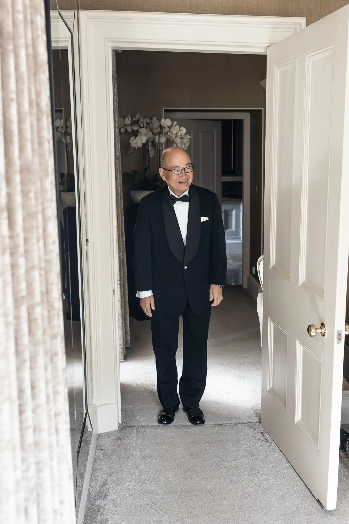 A man in a black tuxedo stands in a doorway, smiling. The room has a mirror, curtain, and visible details like a door handle and a plant in the background.