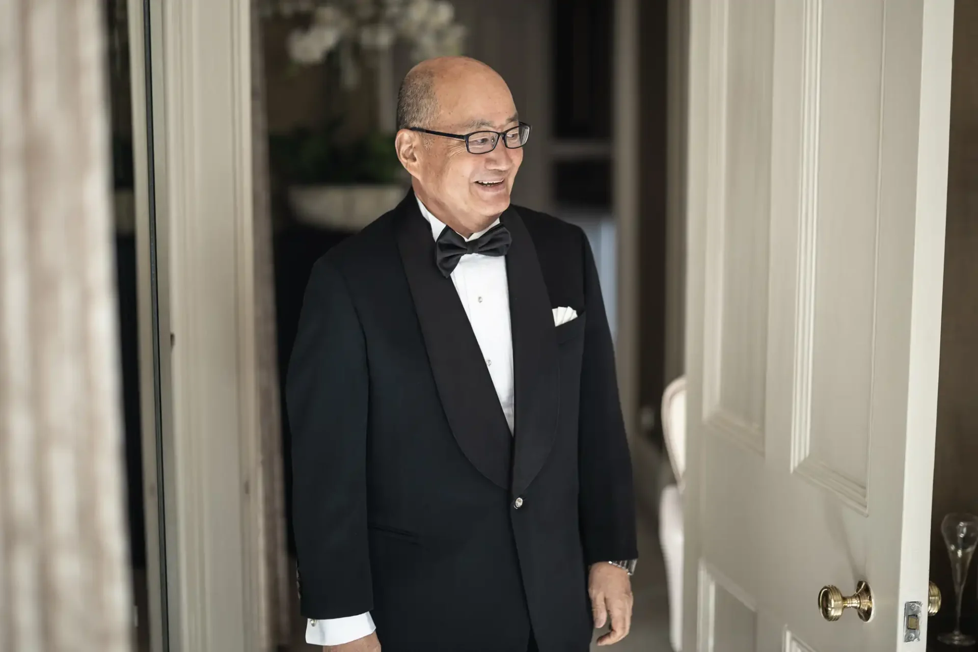 Man in a tuxedo, wearing glasses, smiling and standing in a doorway.