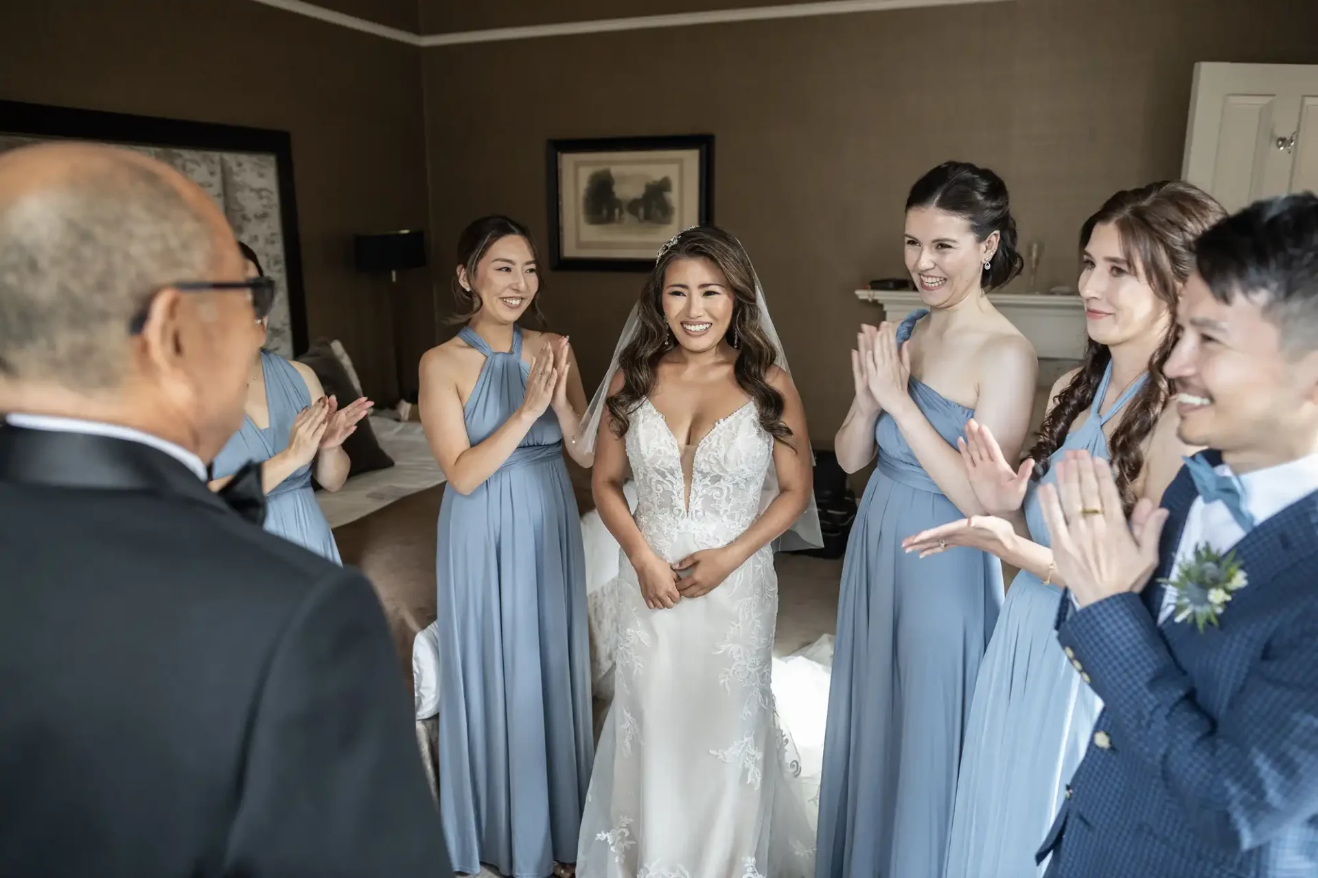 Bride in a white gown, surrounded by four people in matching blue dresses, smiles as they clap in a warmly lit room.