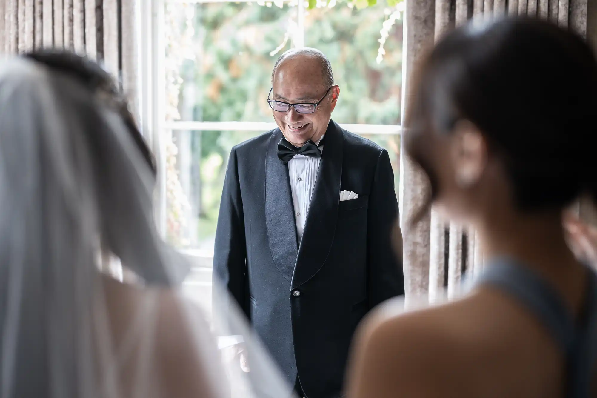 An older man in a tuxedo smiles while standing in front of two blurred figures, one in a wedding dress, by a sunlit window with curtains.