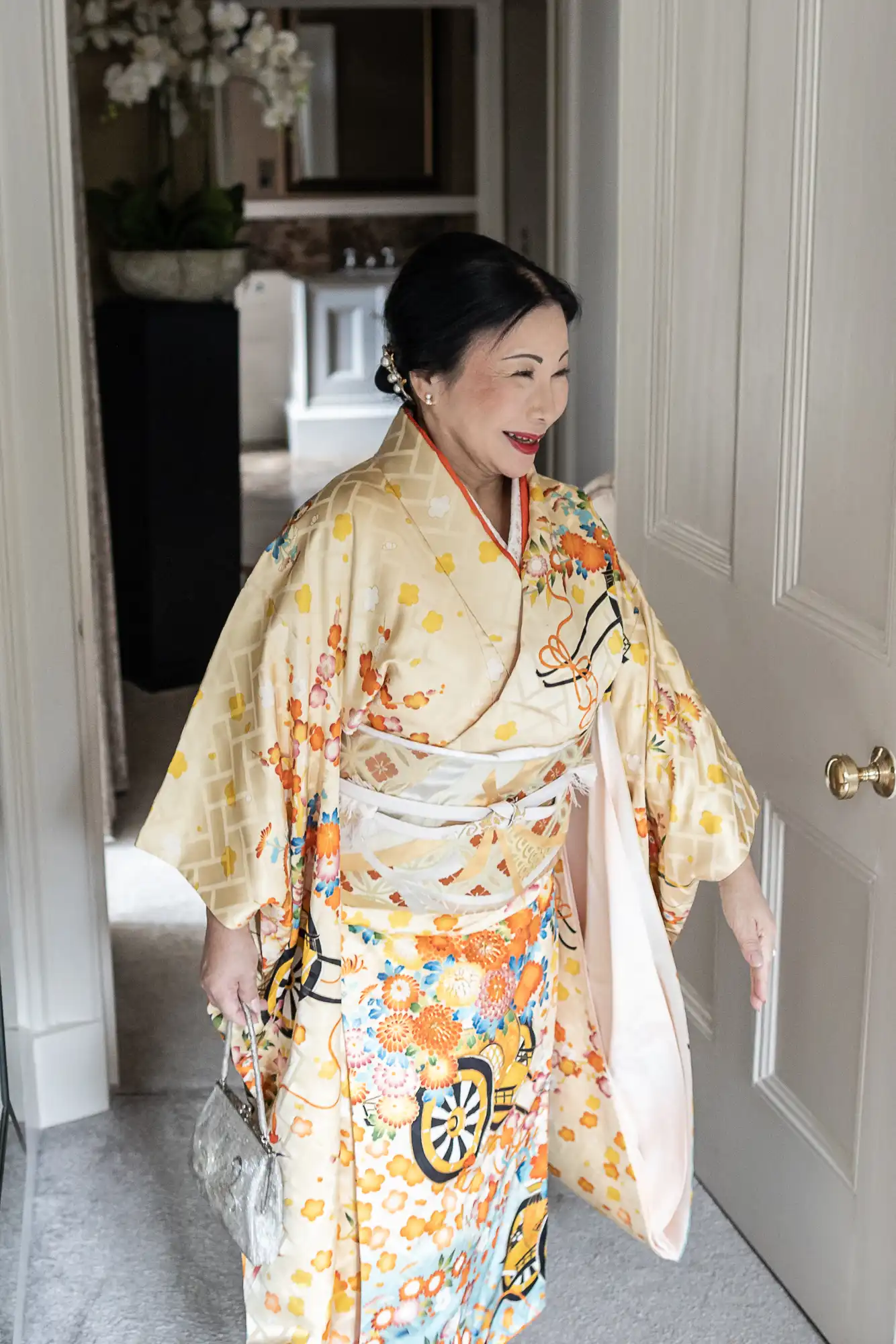 A woman in a colorful kimono stands indoors, holding a small silver handbag. She is near a white door, smiling, with her hair styled neatly.