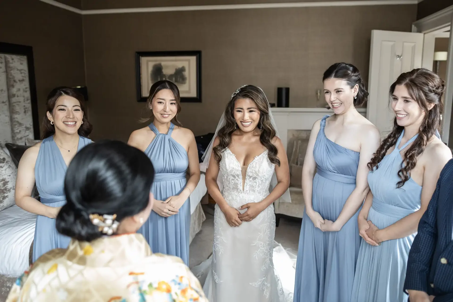 A bride in a white dress stands smiling among five bridesmaids in light blue dresses inside a room with beige walls.