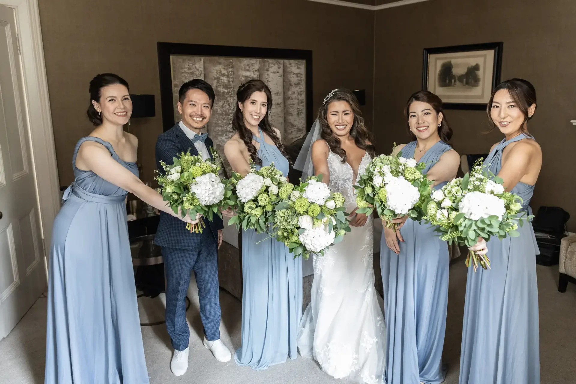 Bride in a white dress poses with five bridesmaids in blue dresses, each holding a bouquet of white and green flowers. They are indoors, smiling at the camera.