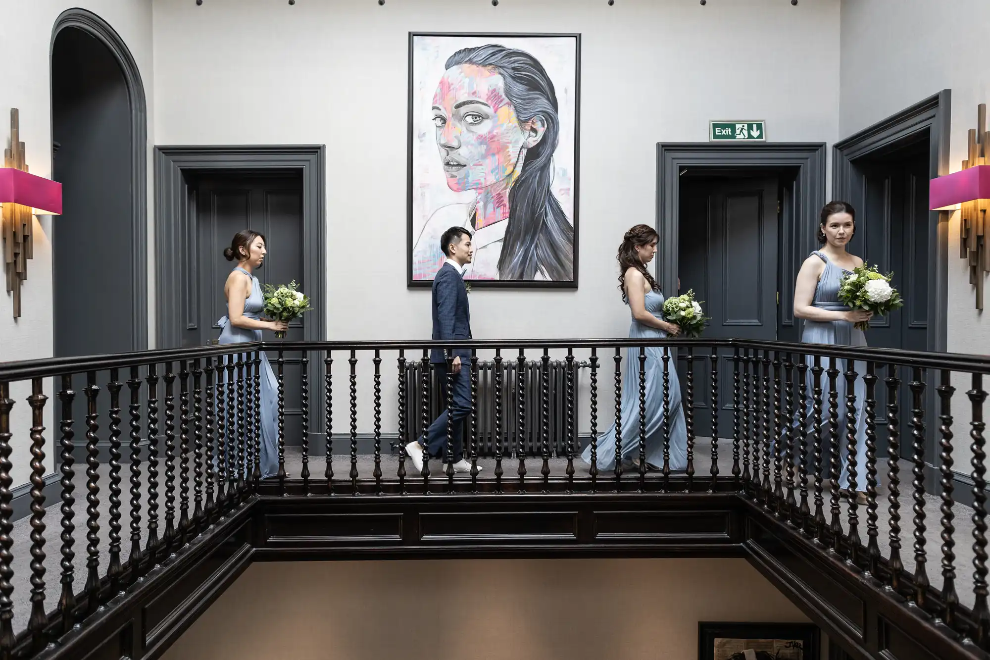 Bridegroom and bridesmaids walk along a hallway with a modern portrait on the wall, carrying bouquets.