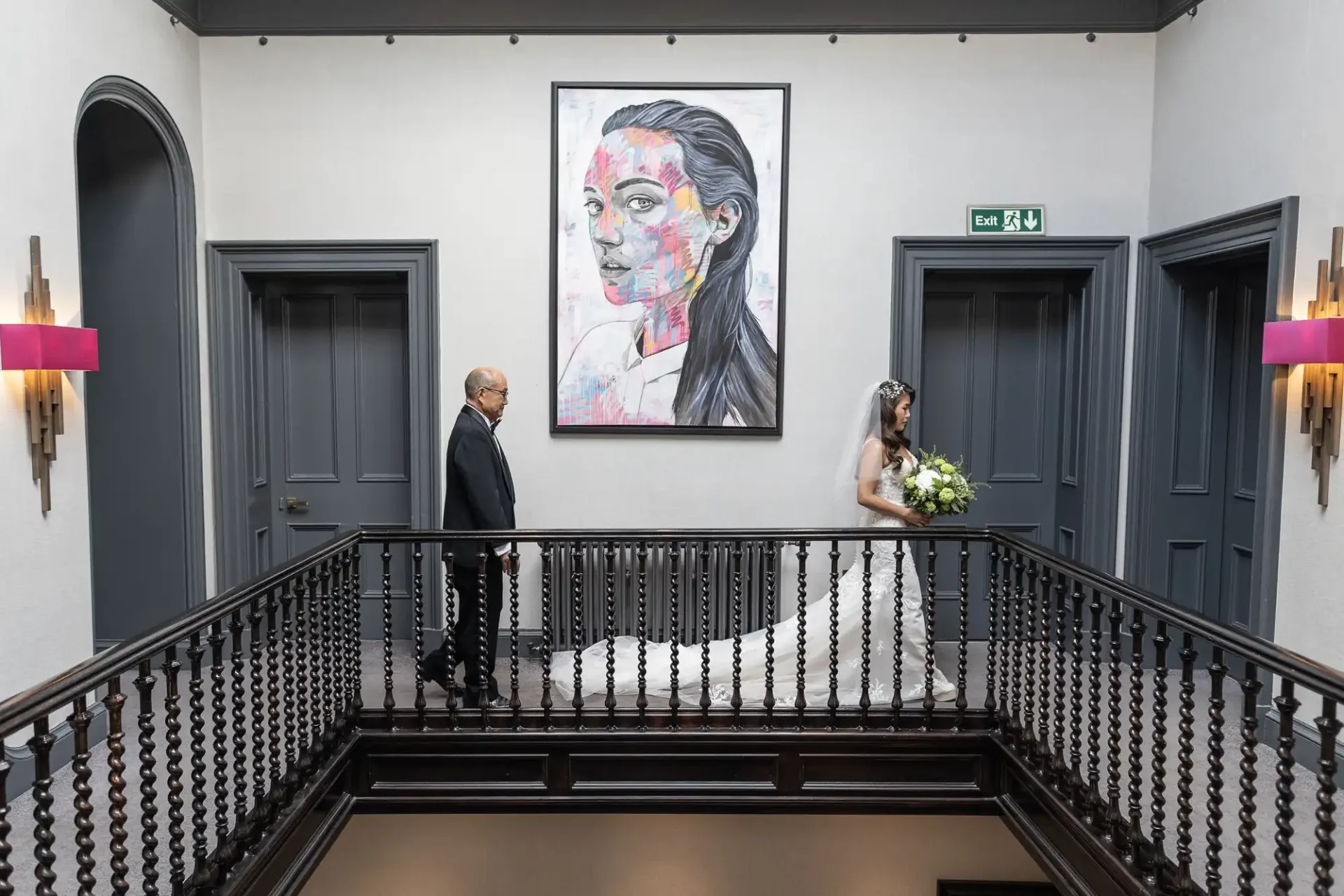 A bride and groom stand on an indoor balcony. The bride is dressed in a white gown with a bouquet, and the groom in a black suit. A large portrait hangs on the wall behind them.