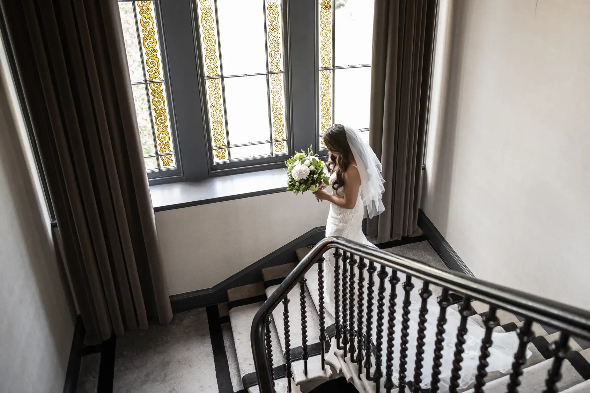 A bride in a white dress and veil descends a staircase, holding a bouquet. She is next to large windows with decorative glass panels.