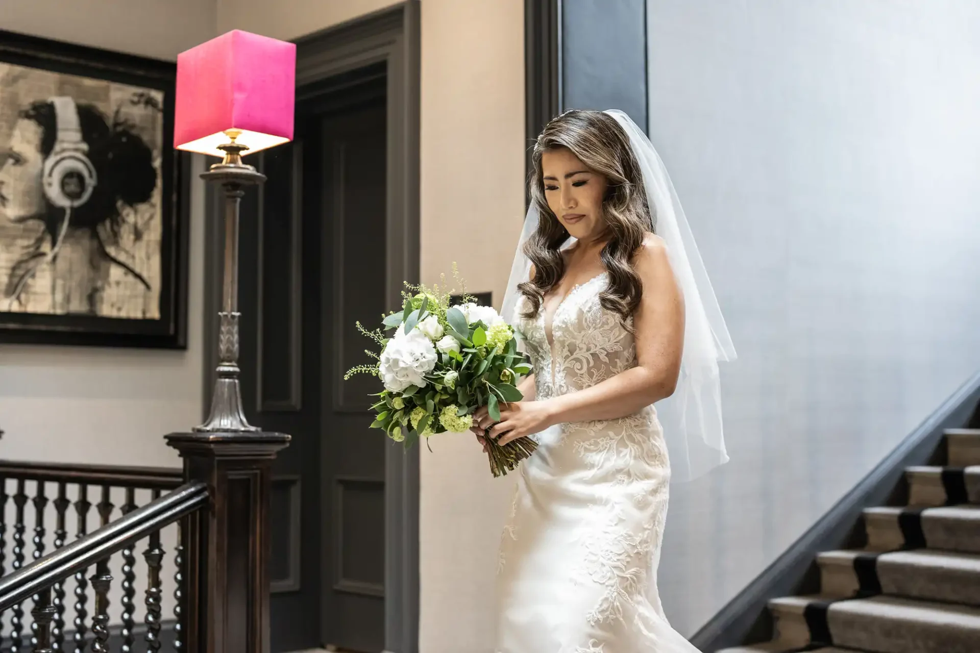 Bride in a white dress and veil descends stairs, holding a bouquet of white and green flowers. A painting of a person with headphones is on the wall.