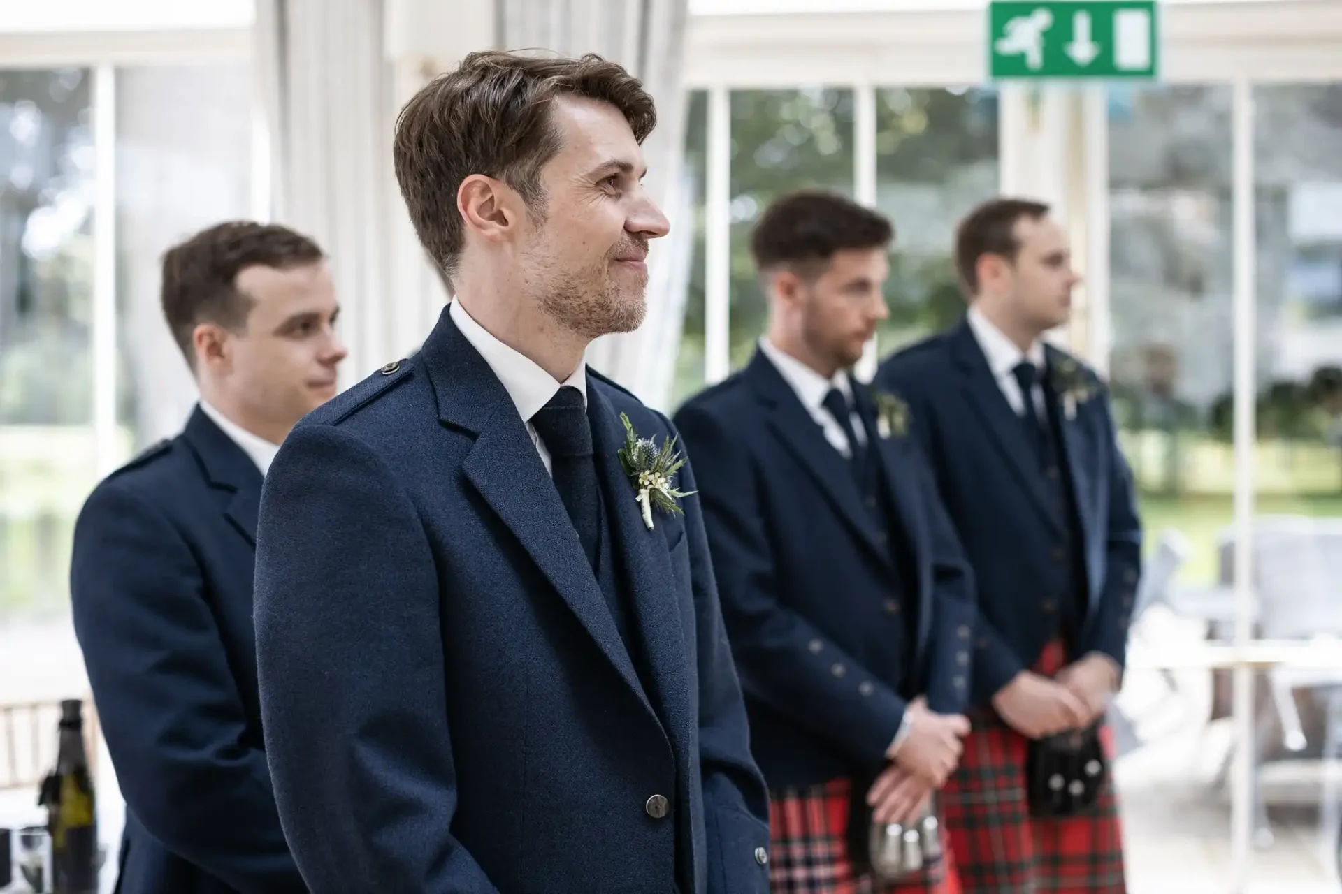 A group of men stand indoors, wearing blue suits and tartan kilts. One man in focus wears a floral boutonniere and smiles slightly.