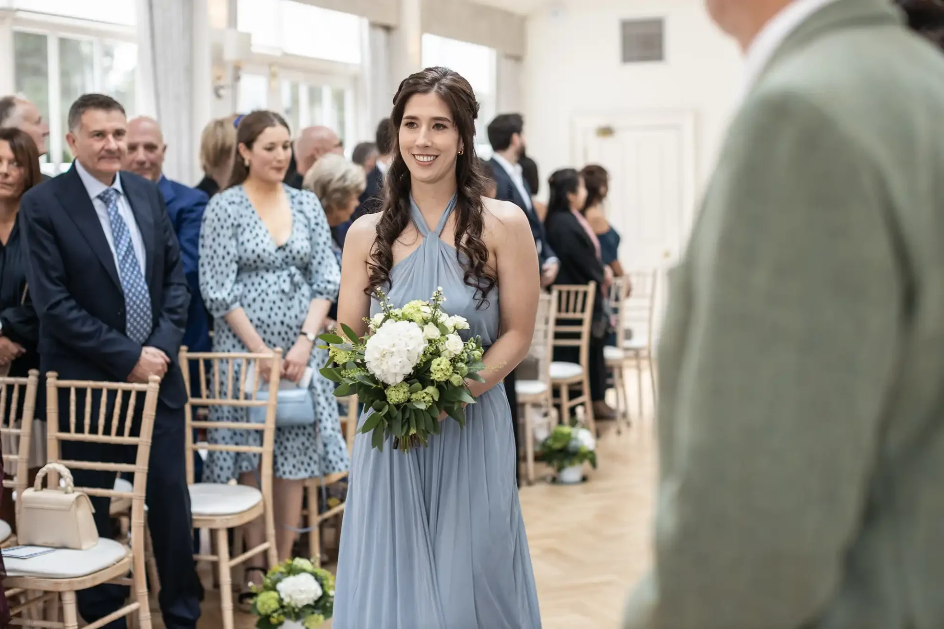 A woman in a blue dress holding a bouquet walks down an aisle in a room filled with seated guests.