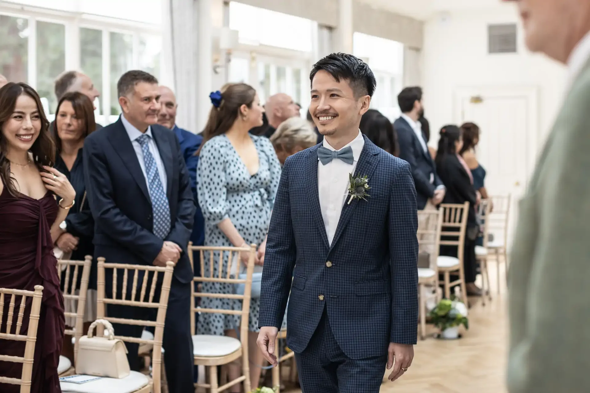 A man in a suit walks down an aisle in a bright room, smiling, with seated and standing guests on either side.