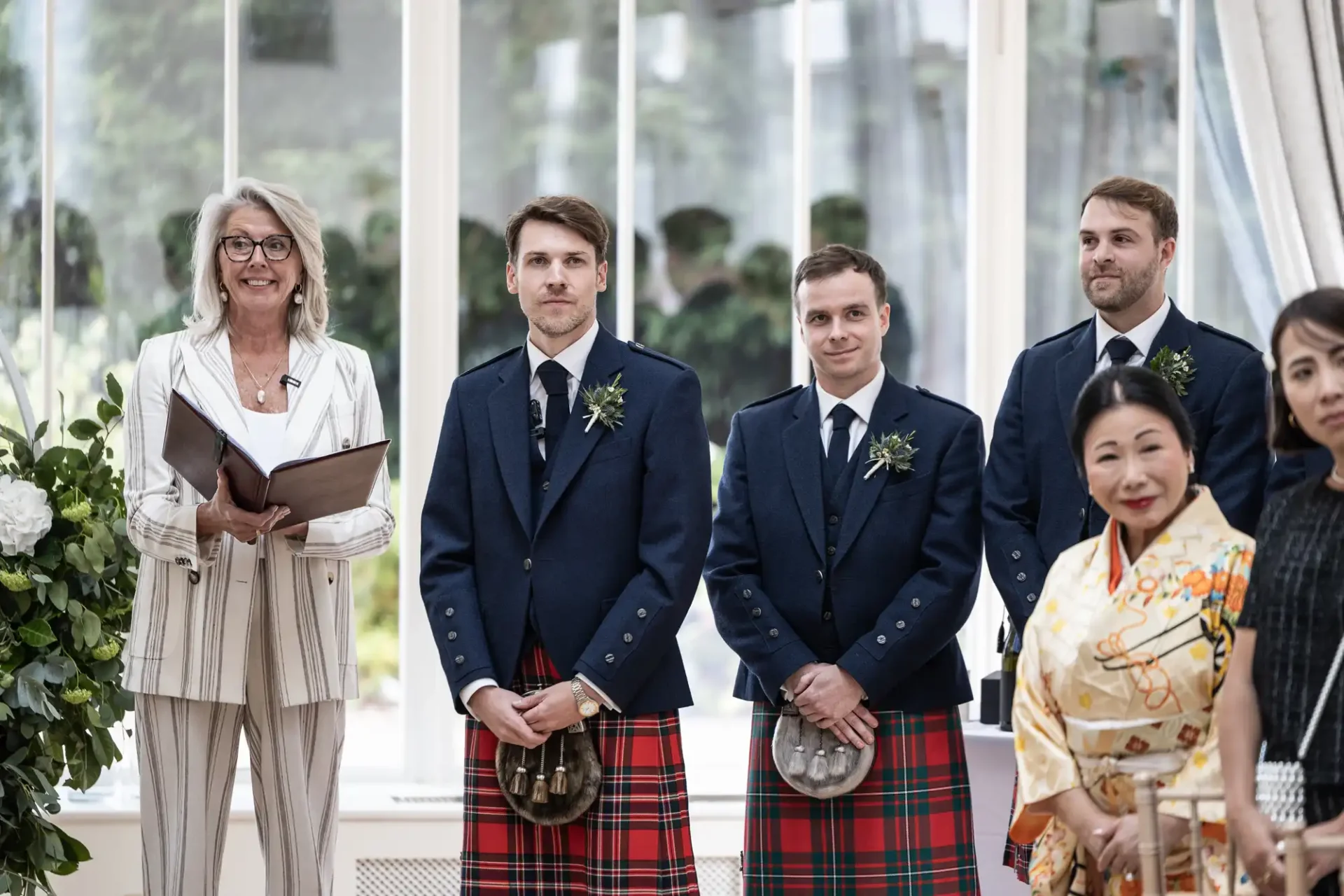 A smiling officiant stands beside three men in kilts and suits. A woman in a kimono is visible in the foreground. They are indoors in front of large windows.