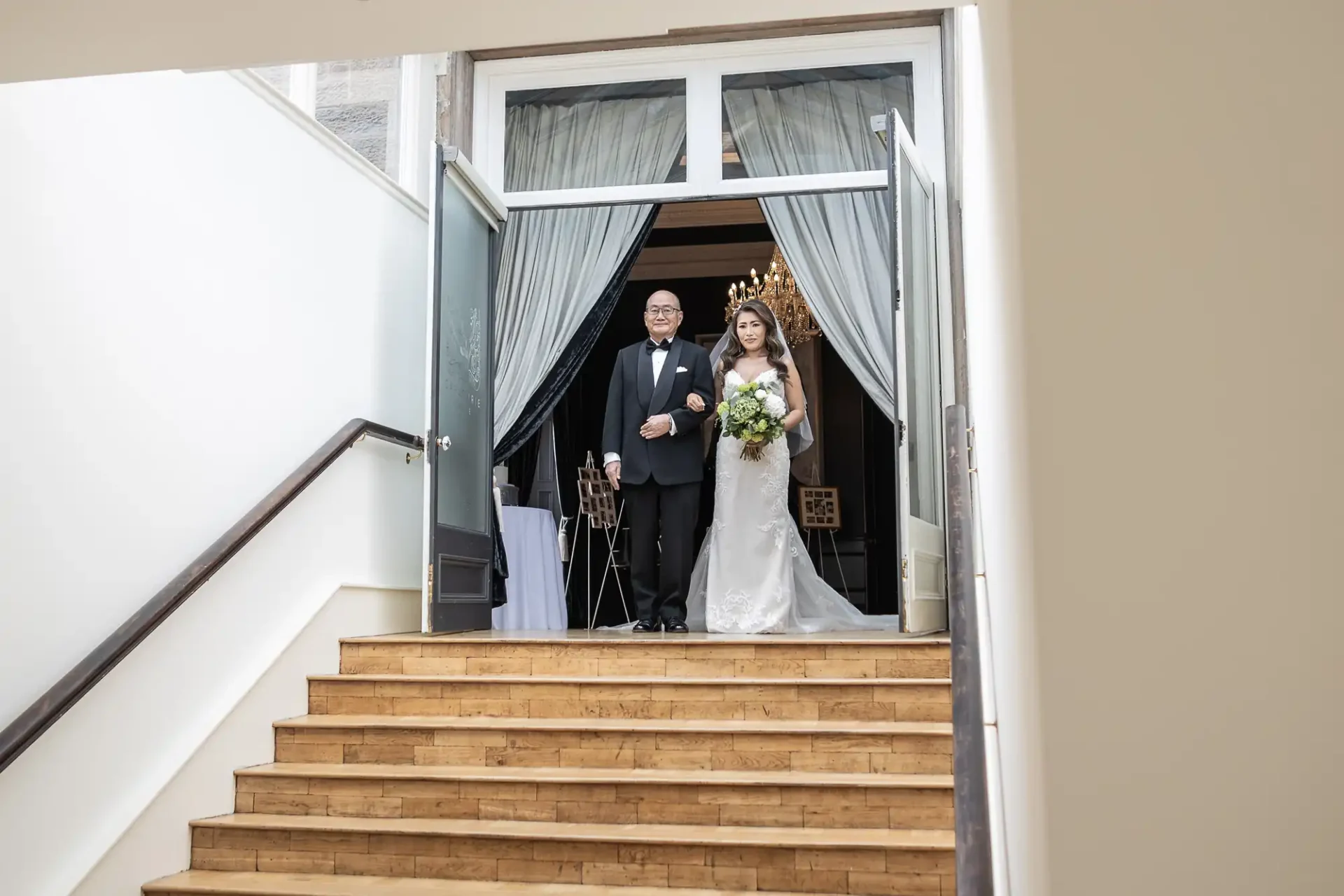 A bride and an older man walk arm in arm down a staircase. The bride wears a white dress and holds a bouquet.