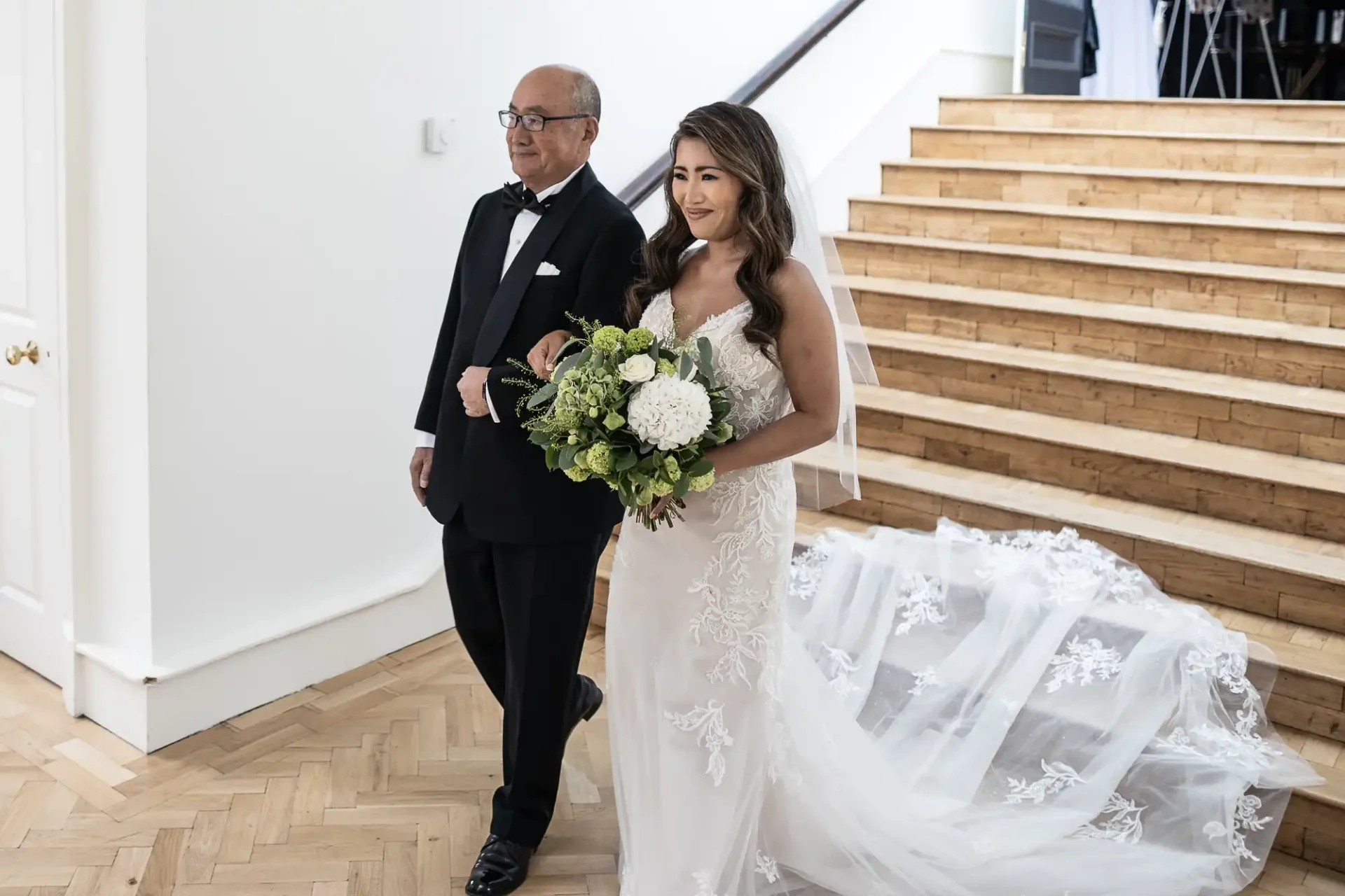 A bride in a white gown walks arm in arm with an older man in a tuxedo, holding a bouquet with white and green flowers, near wooden stairs indoors.