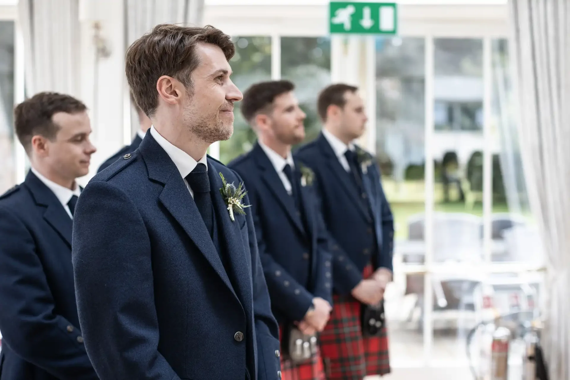 Four men stand indoors in formal attire with navy jackets. One wears a kilt. They are looking in the same direction.