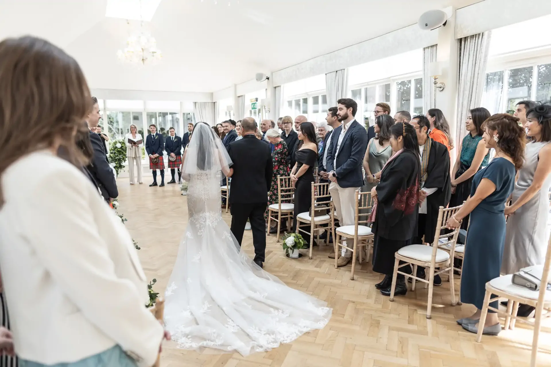 Bride and groom stand at an indoor wedding ceremony surrounded by guests seated and standing. The room is bright with natural light and chandeliers.