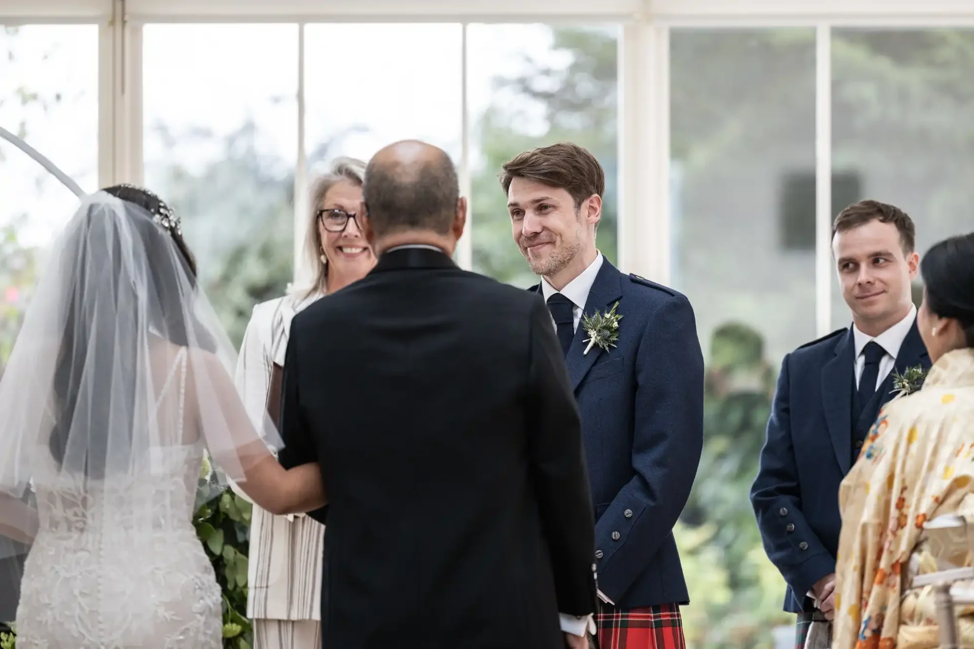 A groom in a blue jacket and red tartan kilt stands beside a man in blue, facing a bride in a white dress and veil, with an officiant and a guest nearby, in a bright room.