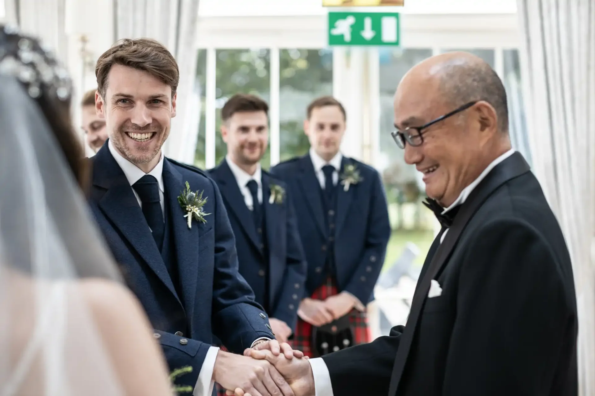 A groom in a blue suit shakes hands with an older man in a tuxedo at a wedding ceremony. Two groomsmen in the background watch the scene.