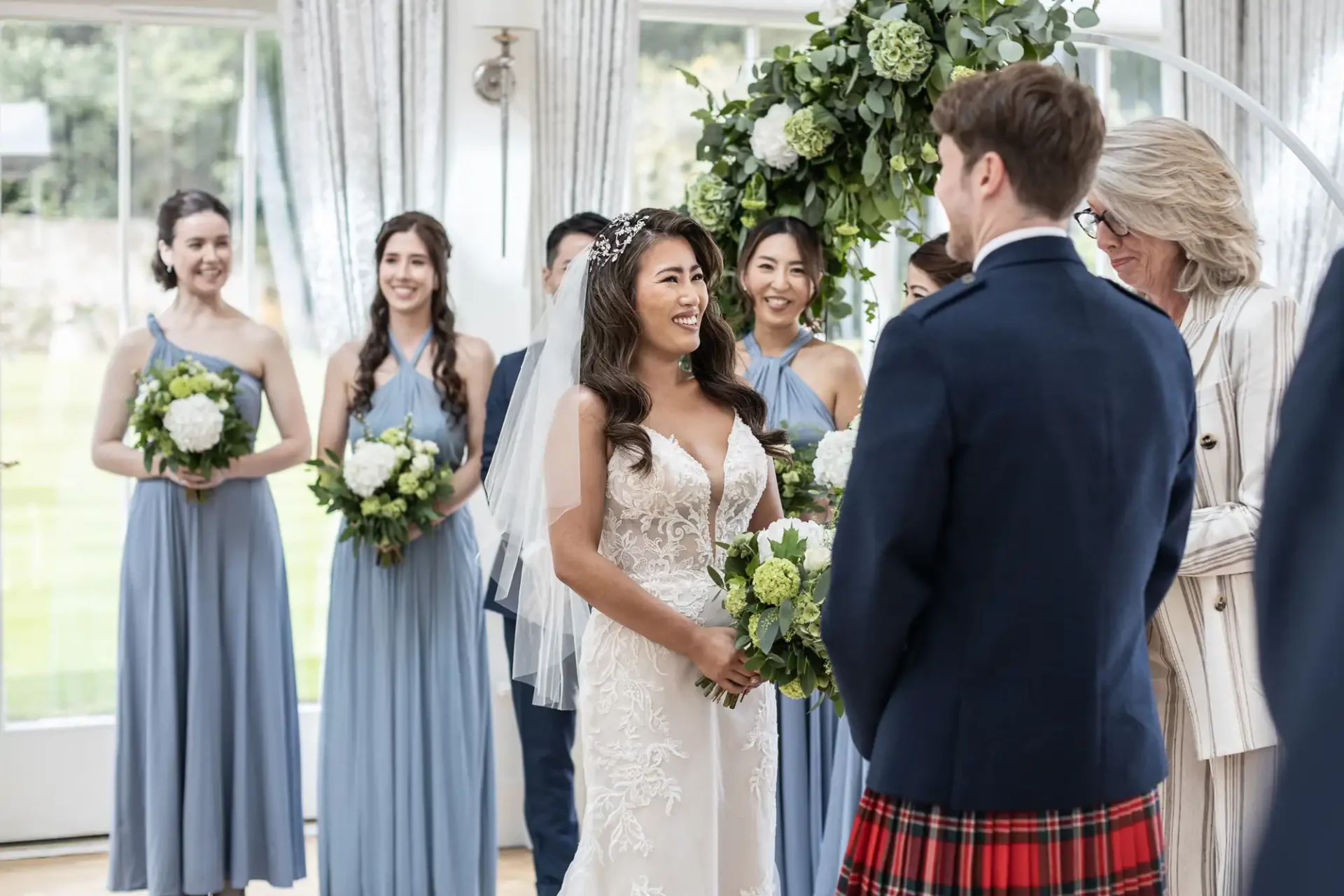 A bride and groom stand with an officiant during a wedding ceremony. The bride smiles in a white gown with a veil, and bridesmaids in blue dresses hold bouquets.