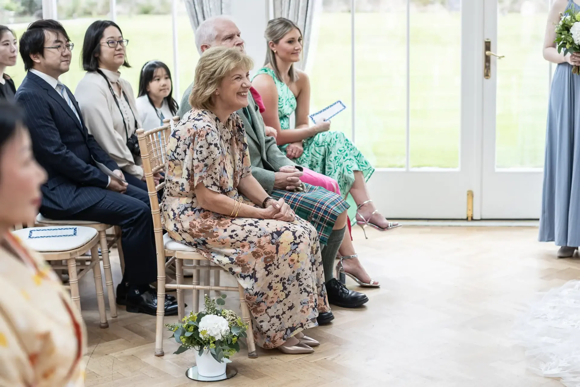 People seated at a wedding ceremony, some dressed in formal attire, with chairs arranged near a large window.