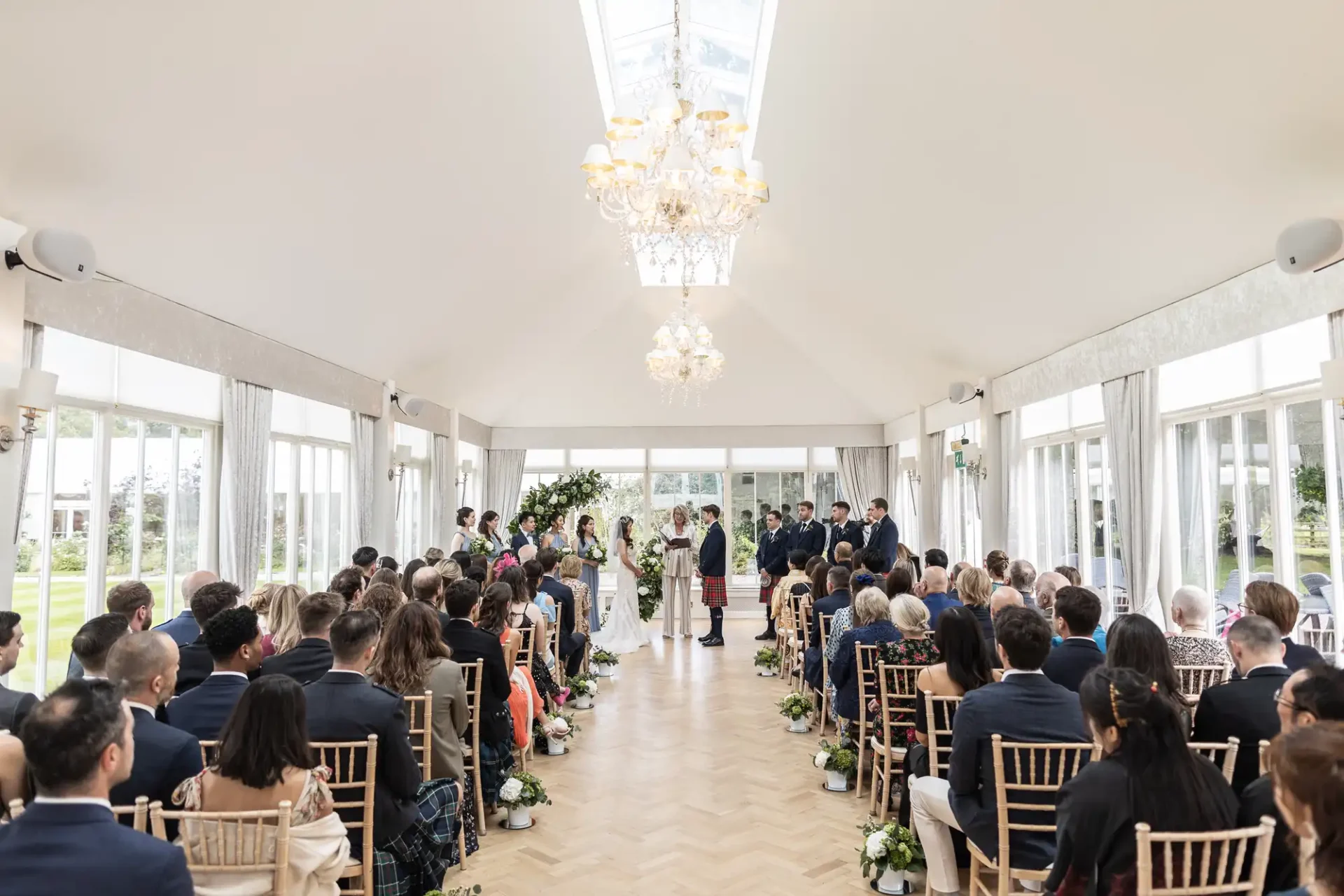 A wedding ceremony taking place indoors, with guests seated on either side of a central aisle. The couple is standing at the front, surrounded by attendants, under a decorative arch.