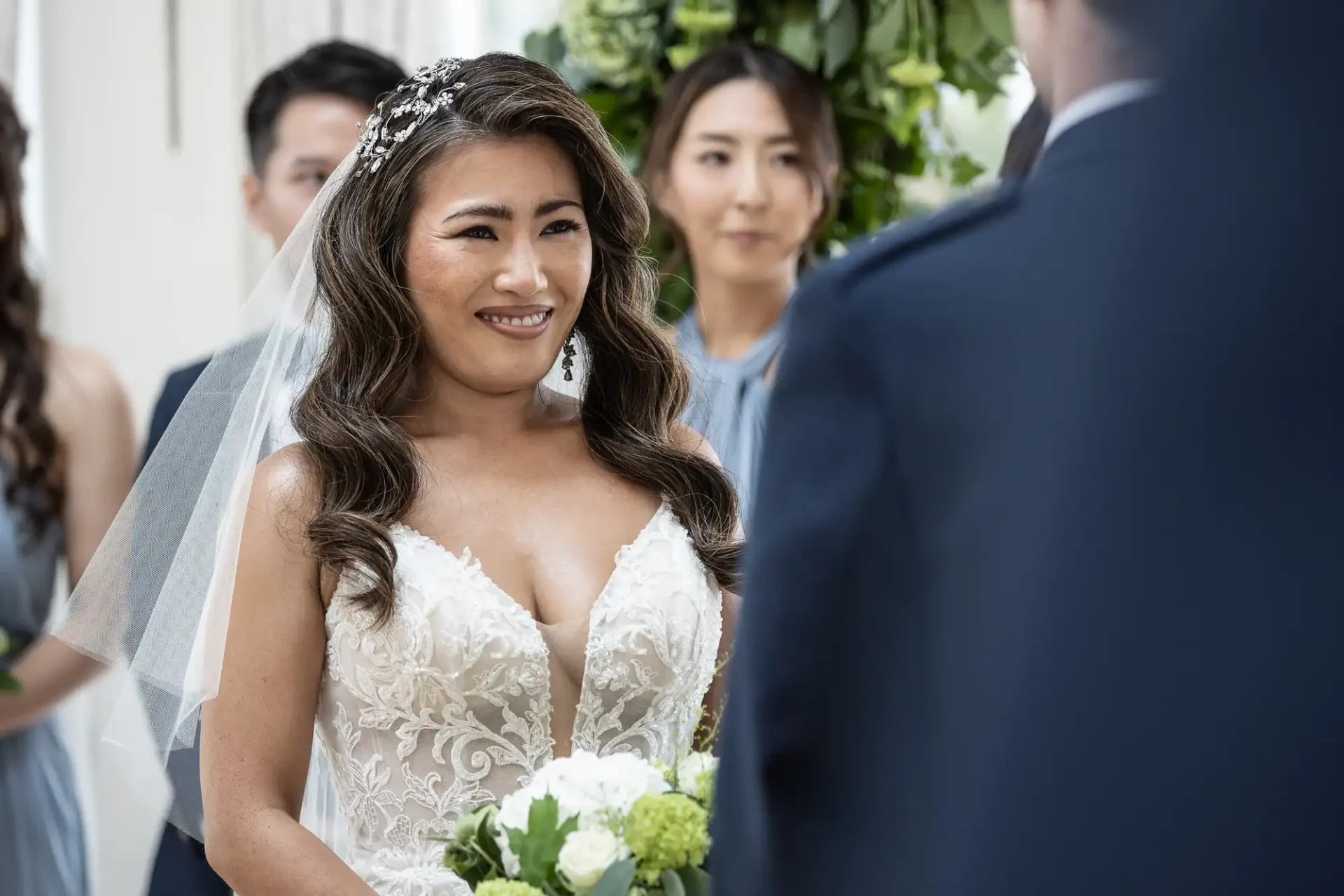 Bride in a white dress and veil smiling at a groom during a wedding ceremony, with bridesmaids in the background.