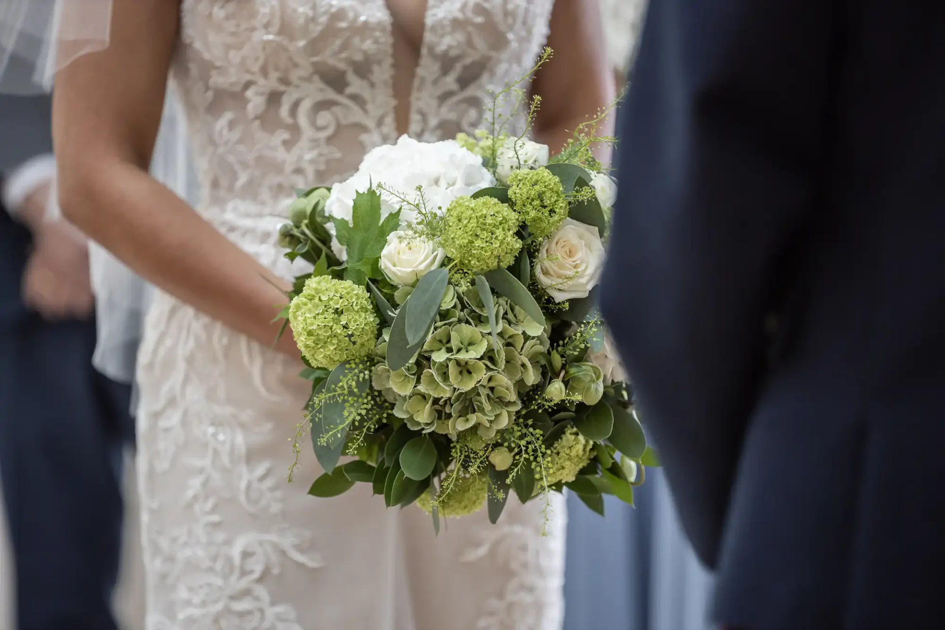 Bride in a lace wedding dress holding a bouquet of white and green flowers, standing beside a person in a dark suit.