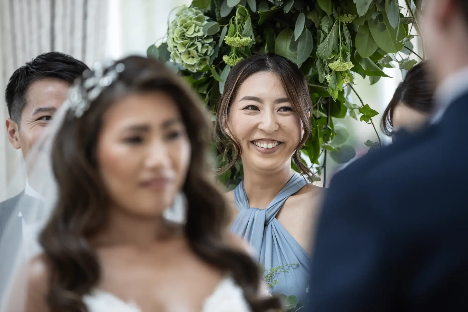 A woman smiles at a wedding ceremony, standing among green floral arrangements.