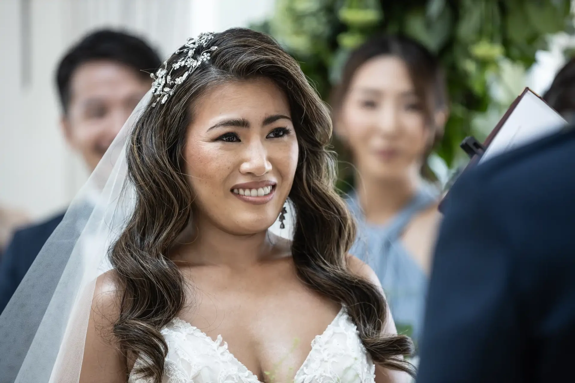 Bride in a white dress and veil, smiling, with people in the background during a ceremony.