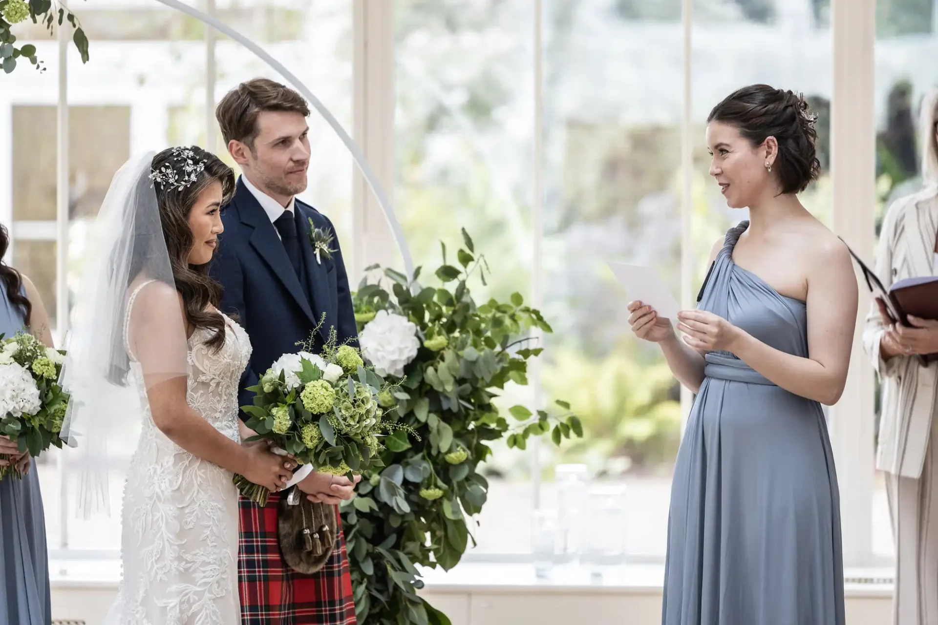 A bride and groom stand facing a bridesmaid who is reading from a paper. The bride holds a bouquet, and the groom wears a flower boutonniere. The setting is bright and decorated with greenery.