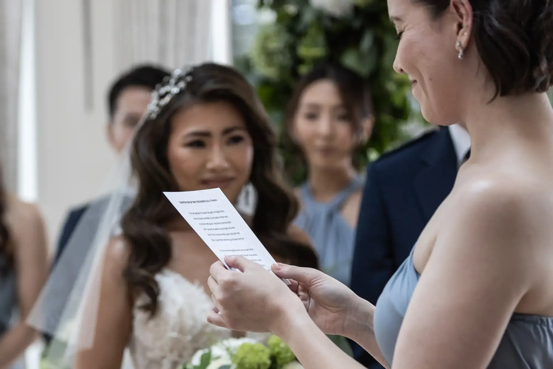 A person reads from a paper during a wedding ceremony, with the bride and other wedding party members in the background.