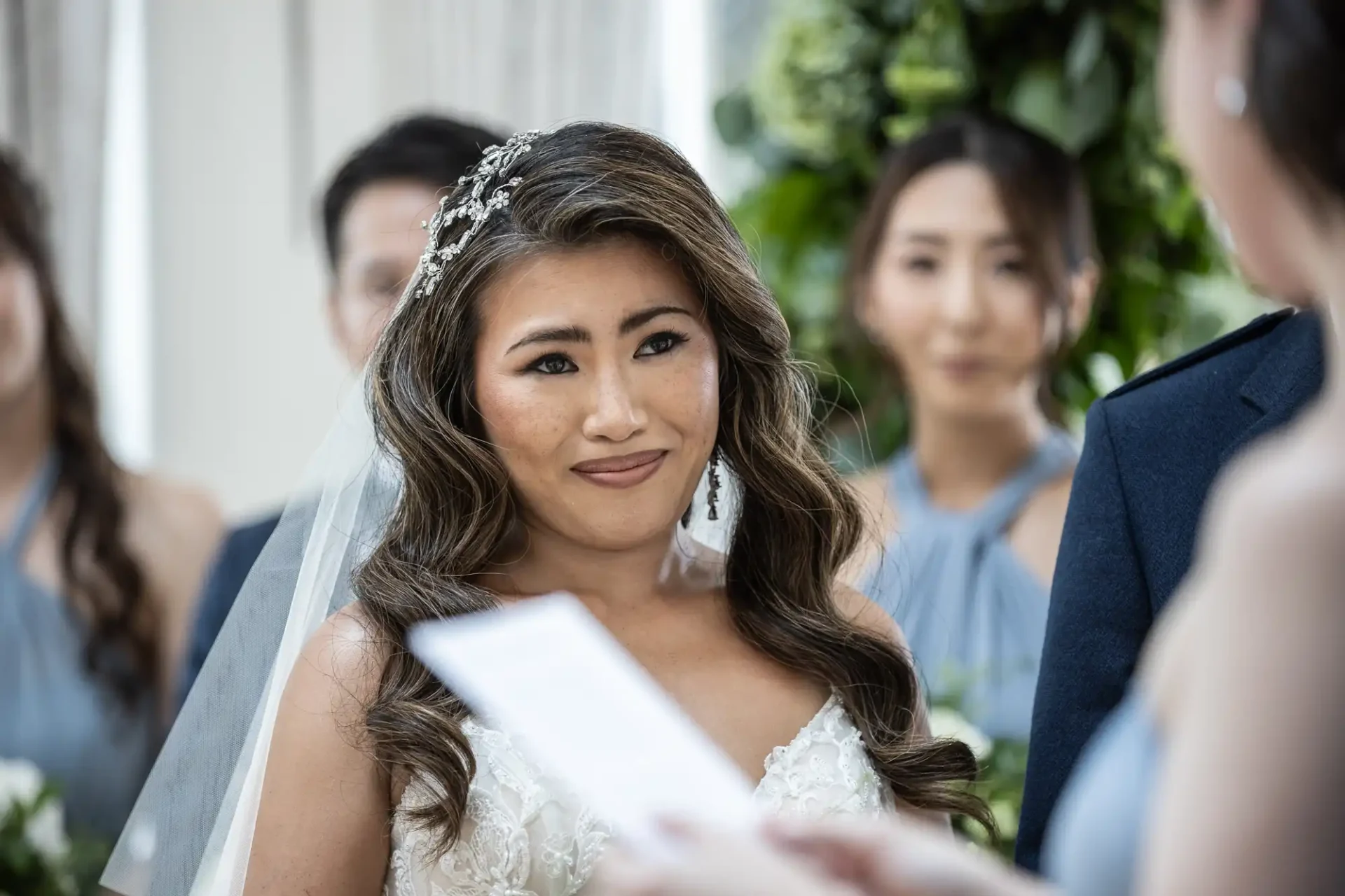 A bride with long wavy hair and a decorative hairpiece looks at someone holding a paper. Bridesmaids in blue dresses are in the background, with greenery visible.