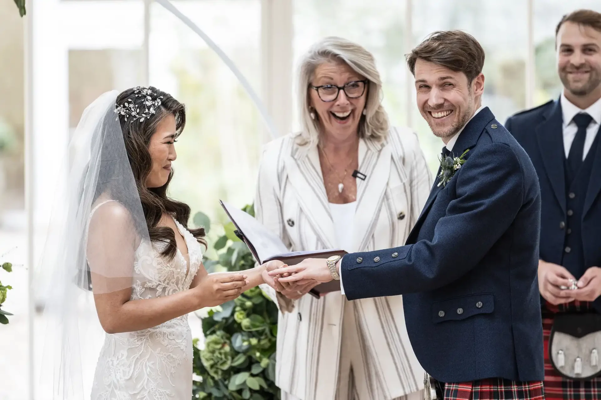 A bride and groom exchange wedding rings while the officiant smiles broadly. Another person stands in the background.