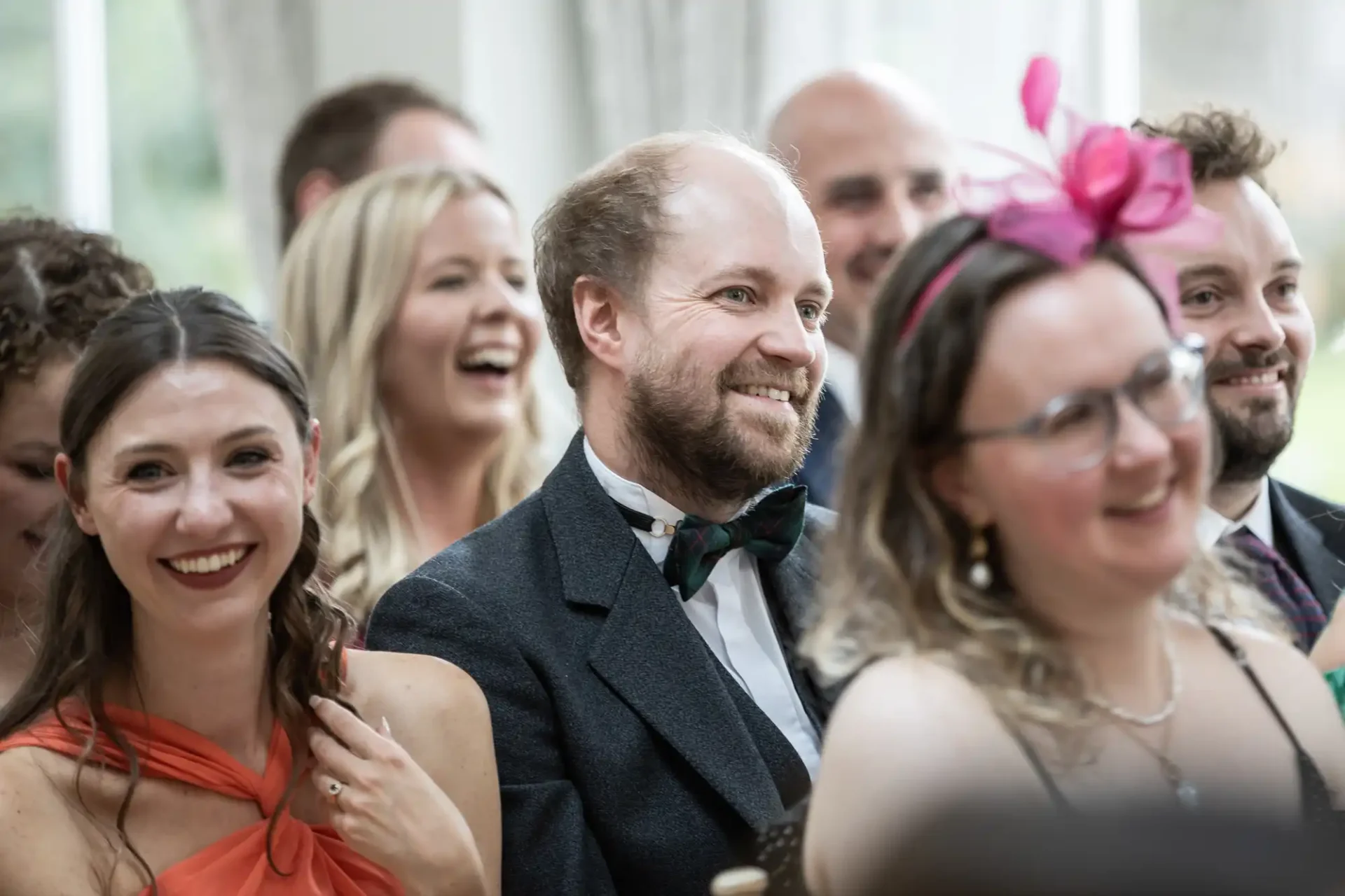 Group of people sitting closely together, smiling and laughing, some wearing formal attire and accessories, in an indoor setting.