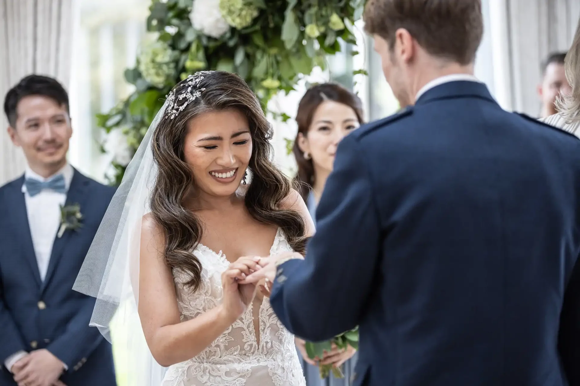 A bride places a ring on a groom's finger during a wedding ceremony. They are surrounded by attendants and greenery.