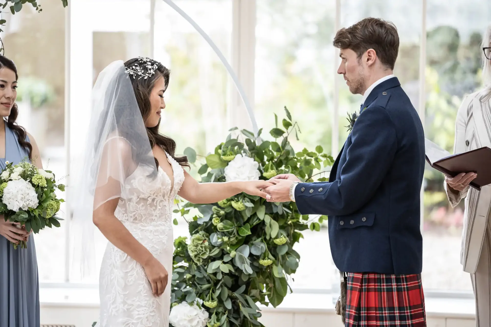 Bride and groom exchange rings at a wedding ceremony. The bride wears a white gown and veil, the groom wears a kilt and blazer. Bridesmaid holds flowers nearby.