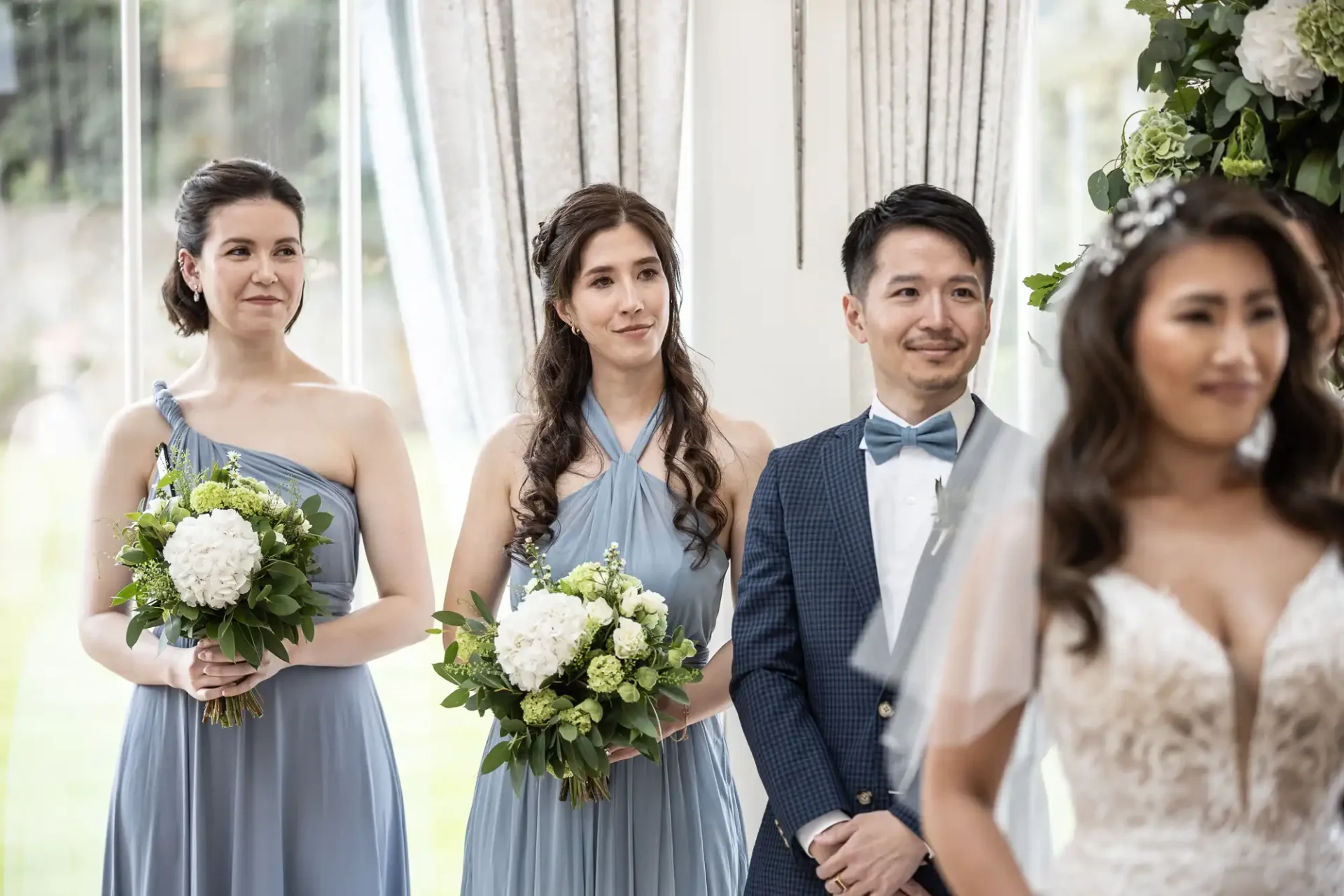 A bride and three attendants at a wedding ceremony. Two women in blue dresses hold white floral bouquets, while a man in a blue suit stands beside them. The focus is on the bride.