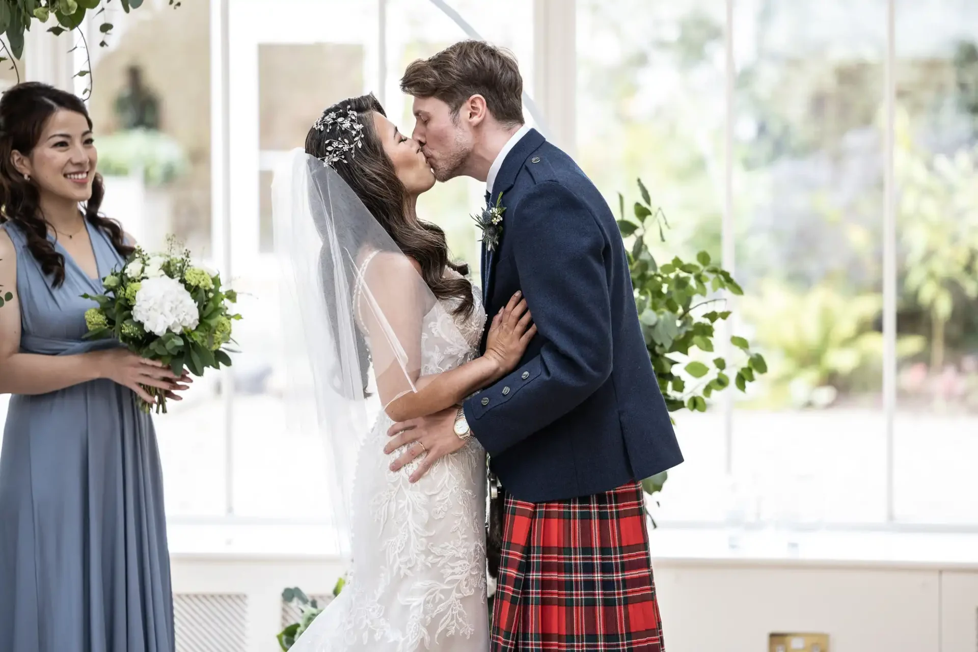 A bride and groom kiss during their wedding ceremony. The bride wears a lace dress and veil, while the groom wears a kilt and jacket. A bridesmaid in blue stands nearby holding flowers.