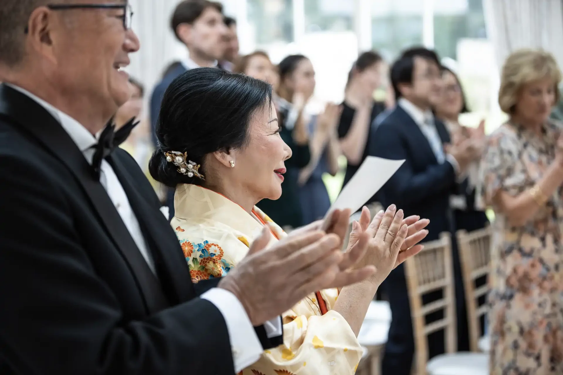 Guests in formal attire clapping at an event; a woman in a yellow floral robe and a man in a tuxedo are in the foreground.