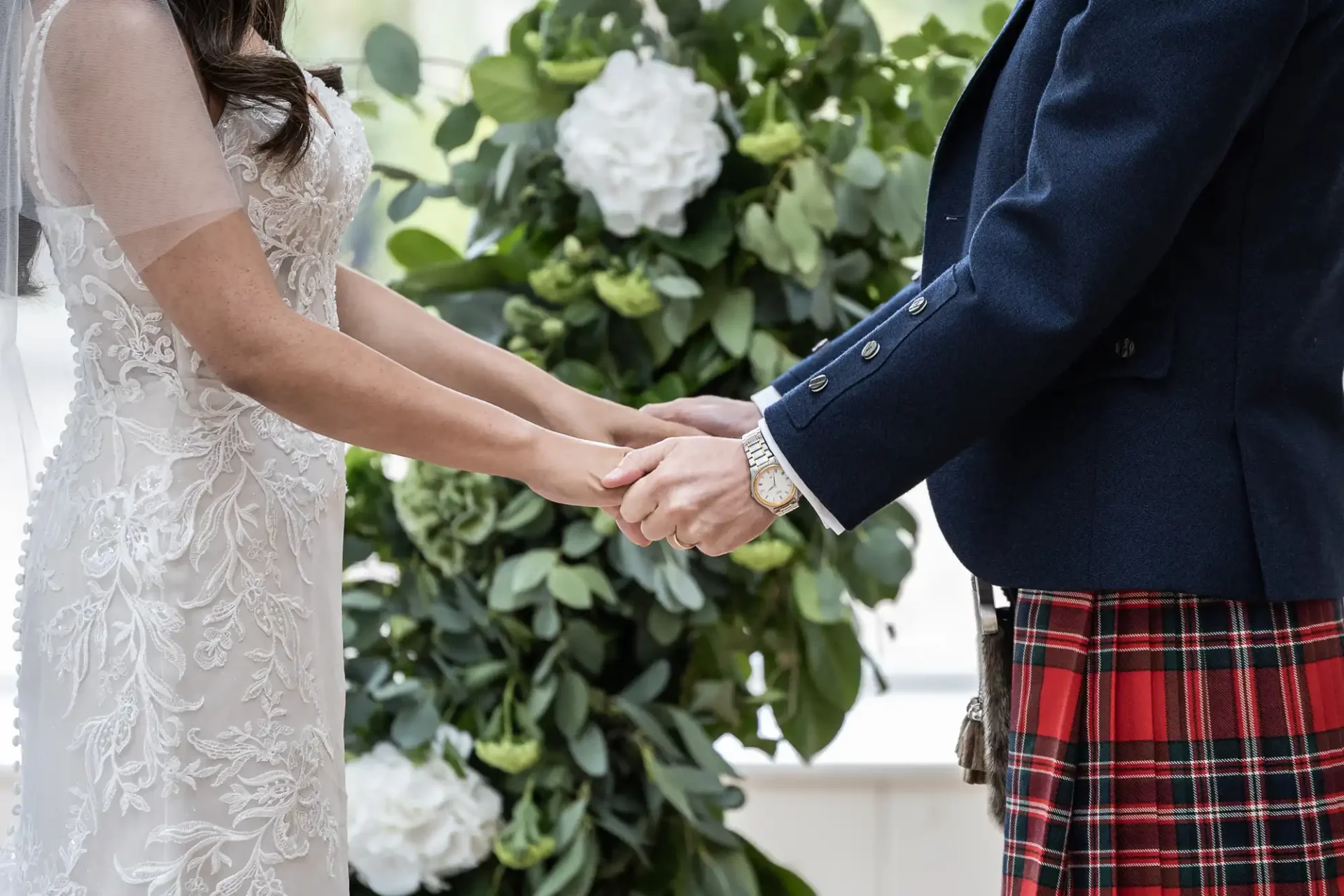 Bride in white lace gown and groom in tartan kilt holding hands in front of greenery.