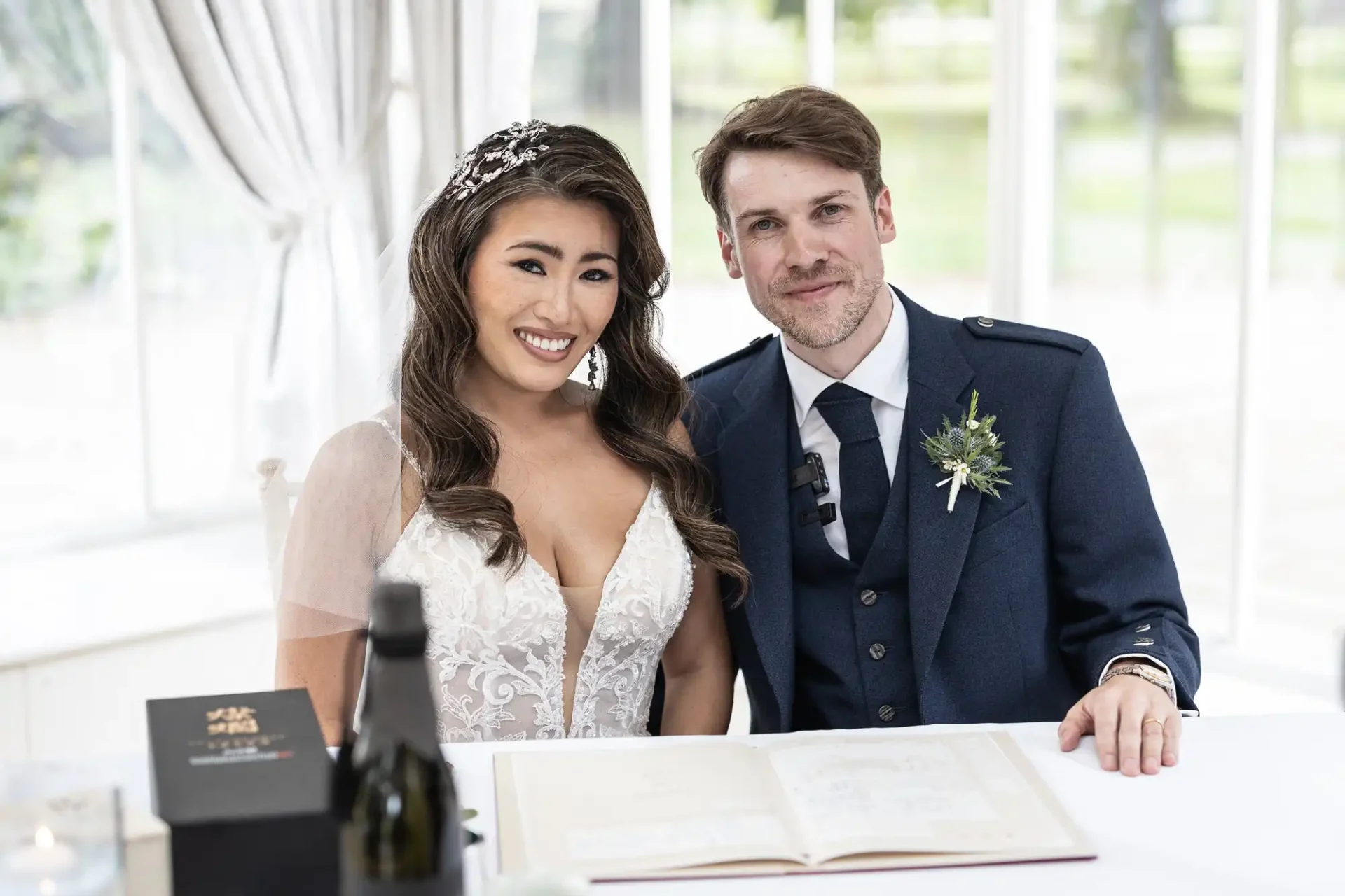 A couple in wedding attire is seated at a table with a book and a bottle. The woman is smiling, wearing a white dress, and the man is in a dark suit with a boutonniere.