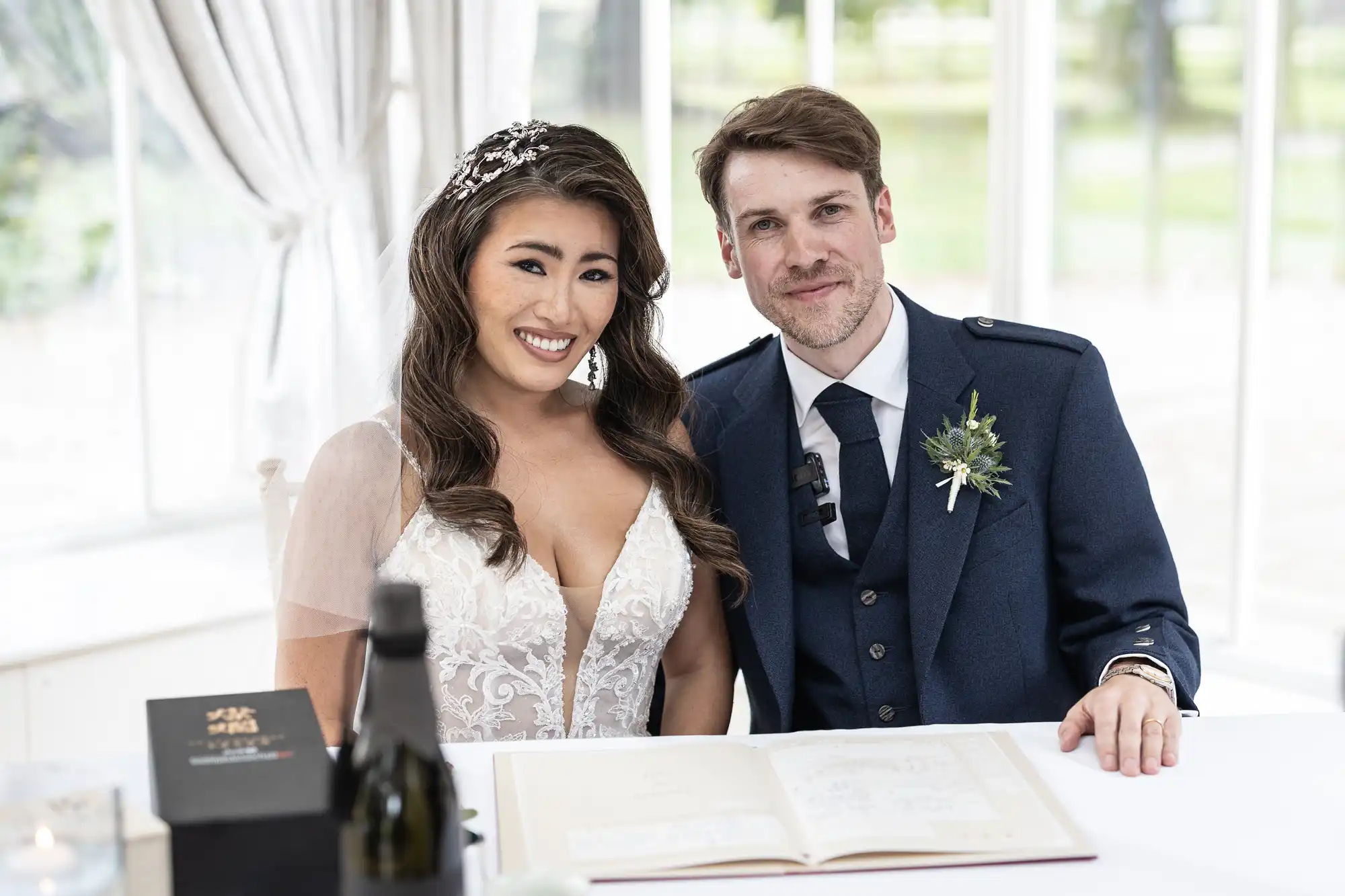 A couple in wedding attire is seated at a table with a book and a bottle. The woman is smiling, wearing a white dress, and the man is in a dark suit with a boutonniere.