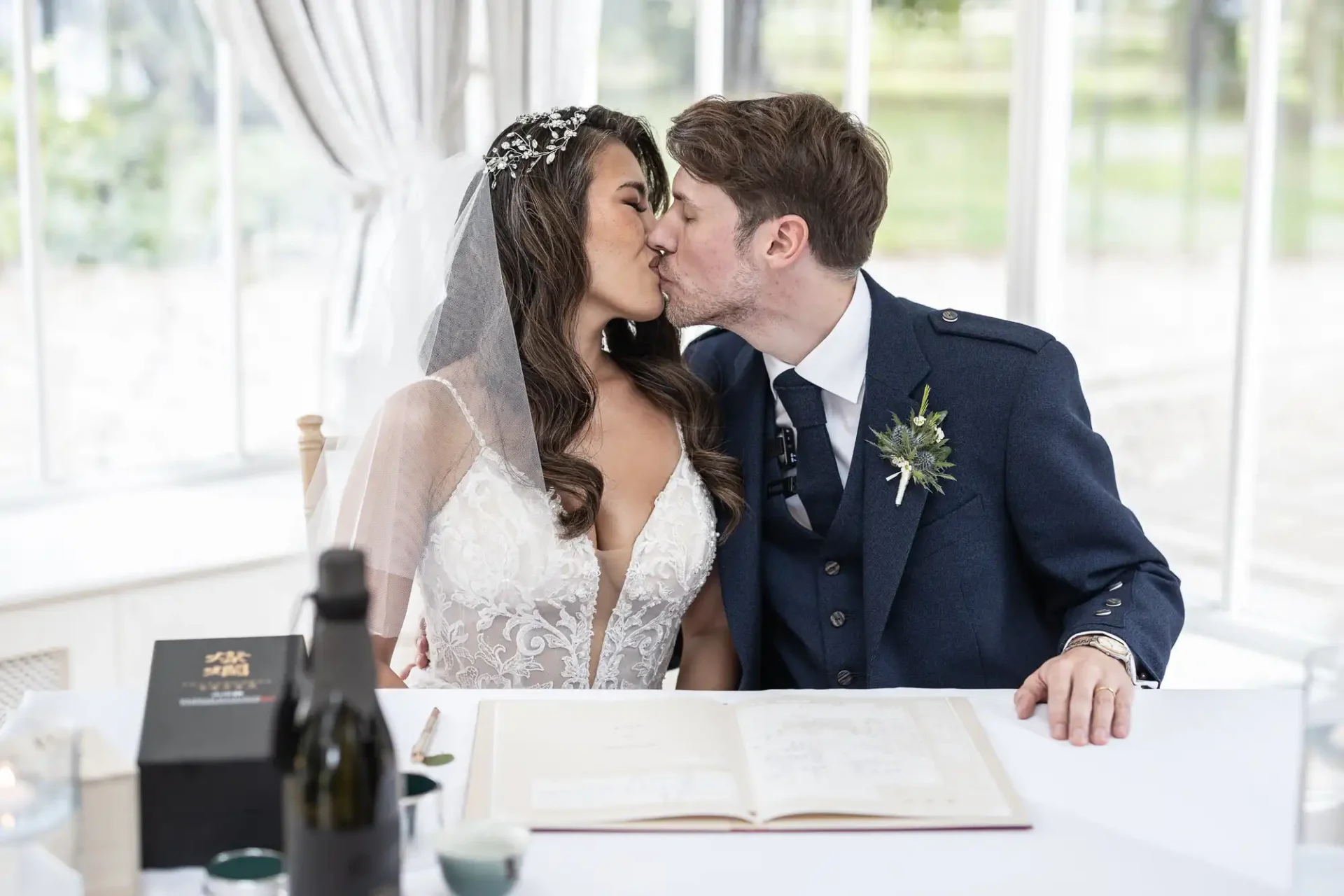 A newlywed couple kisses at a table with a wedding registry book, a pen, and a champagne bottle. The bride wears a veil and floral headpiece; the groom wears a dark suit with a boutonniere.